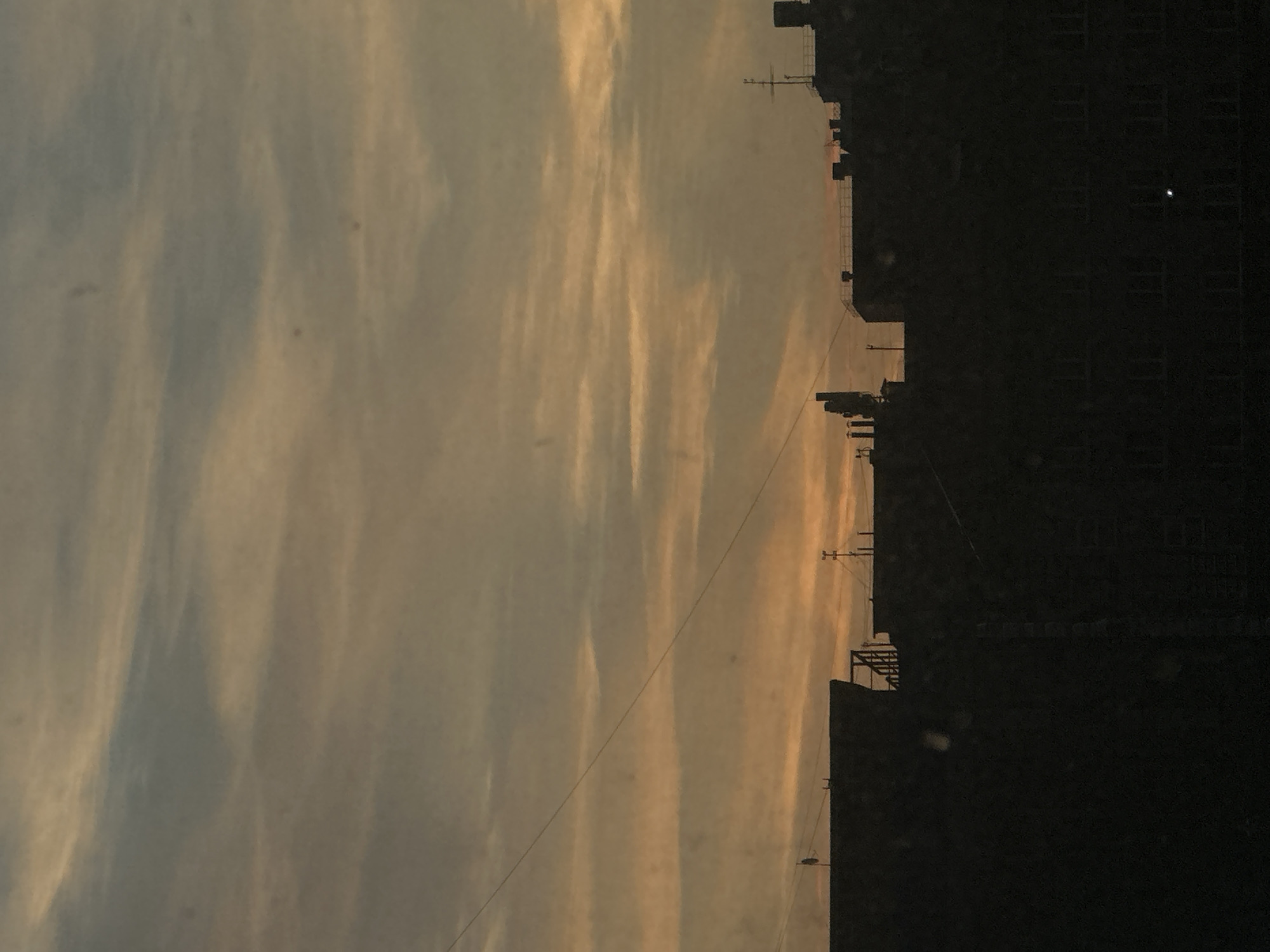 The image depicts a sunset sky over a city, with buildings silhouetted against the sky on the right side of the frame. The sky displays a mix of blue and orange hues, indicative of sunset. A power line cuts diagonally across the sky. Details within the buildings are not clear, but windows and building structures are visible. The location is presumed to be Perm, Russia.