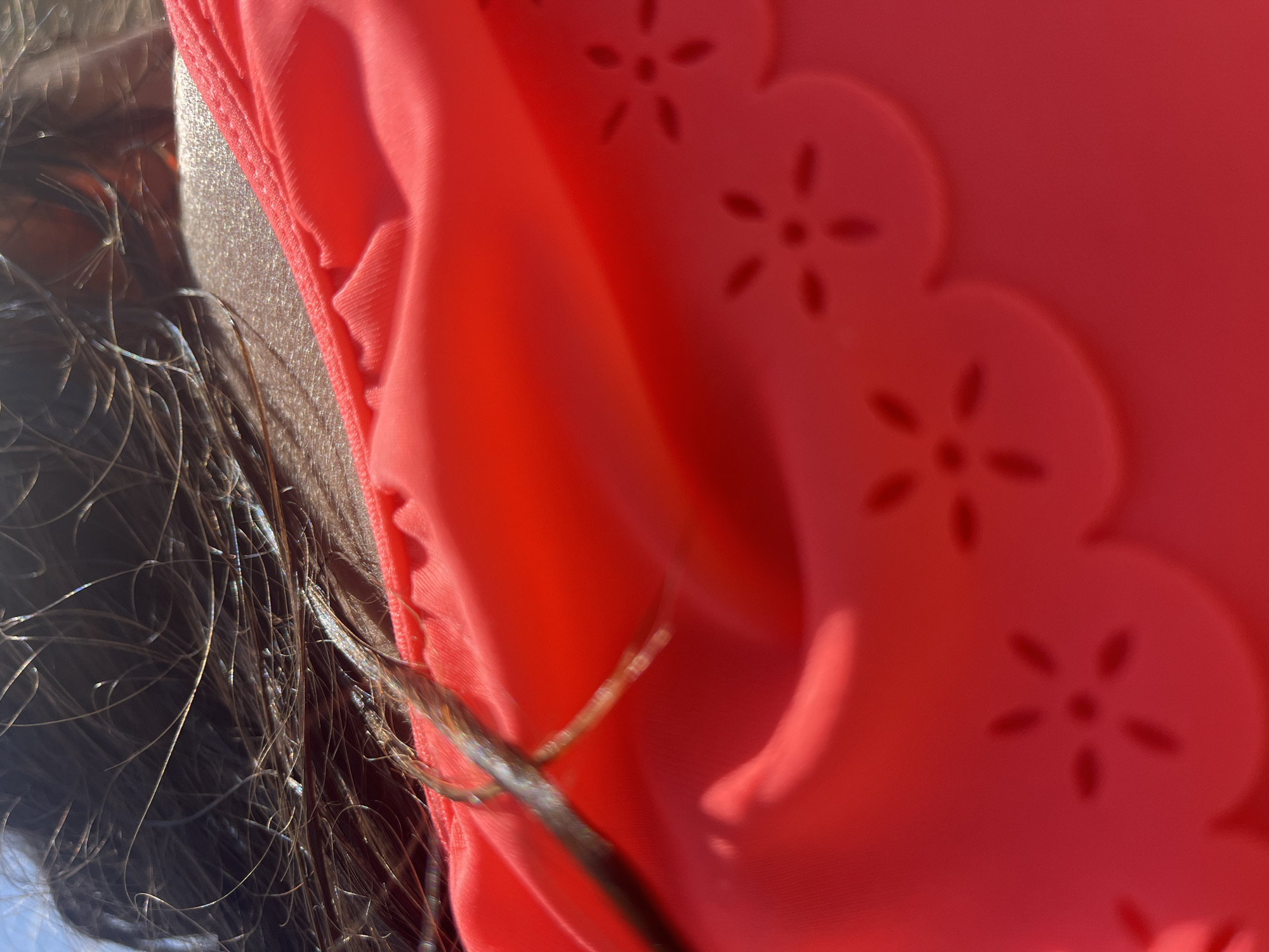 The image shows a close-up of a person's neck and hair, wearing a bright red garment. The hair is dark and appears wet, with some strands reflecting light. The red garment is textured, with a scalloped edge and a subtle floral pattern of cutouts. The lighting suggests it is daytime, possibly outdoors, with sunlight creating highlights on the skin and hair. The scene is abstract and lacks specific environmental context, making it impossible to determine a precise location.