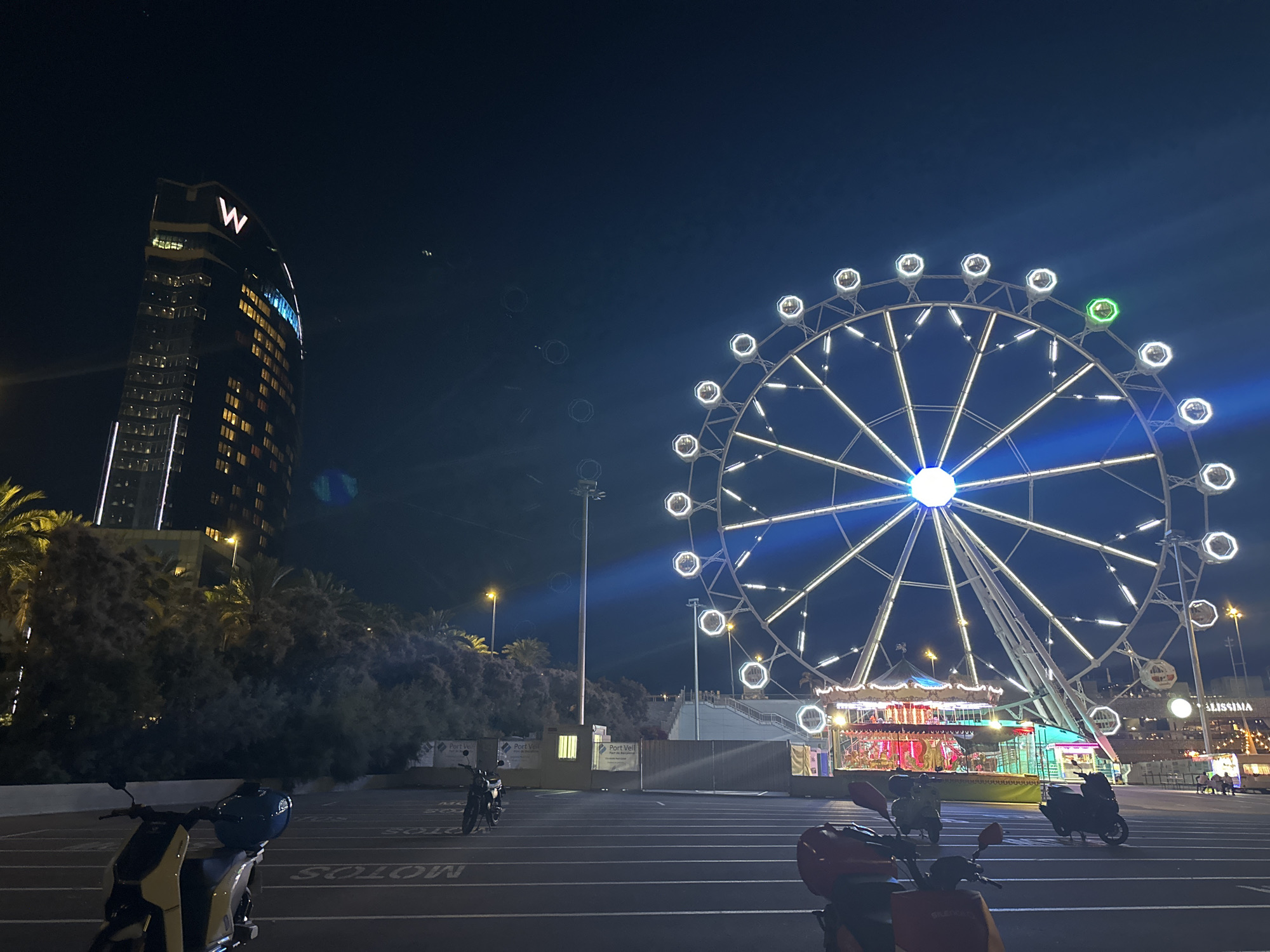 In Barcelona, Spain, at night, a Ferris wheel illuminated with white lights stands prominently in the foreground. To the left of the Ferris wheel, a tall, dark skyscraper with the "W" logo visible at its apex dominates the skyline. The skyscraper features numerous illuminated windows, suggesting it is a hotel or office building. In front of the skyscraper and Ferris wheel, there is a parking lot with several parked motorcycles and scooters. The ground is asphalt with white painted lines indicating parking spaces. Behind the Ferris wheel and to the right, a carousel with colorful lights is visible, along with other amusement park structures and buildings. Trees and vegetation are scattered throughout the scene, particularly around the base of the skyscraper and in the mid-ground. The sky is dark, indicating nighttime, with a few faint lights visible in the distance. The overall atmosphere is that of a lively urban entertainment area at night.
