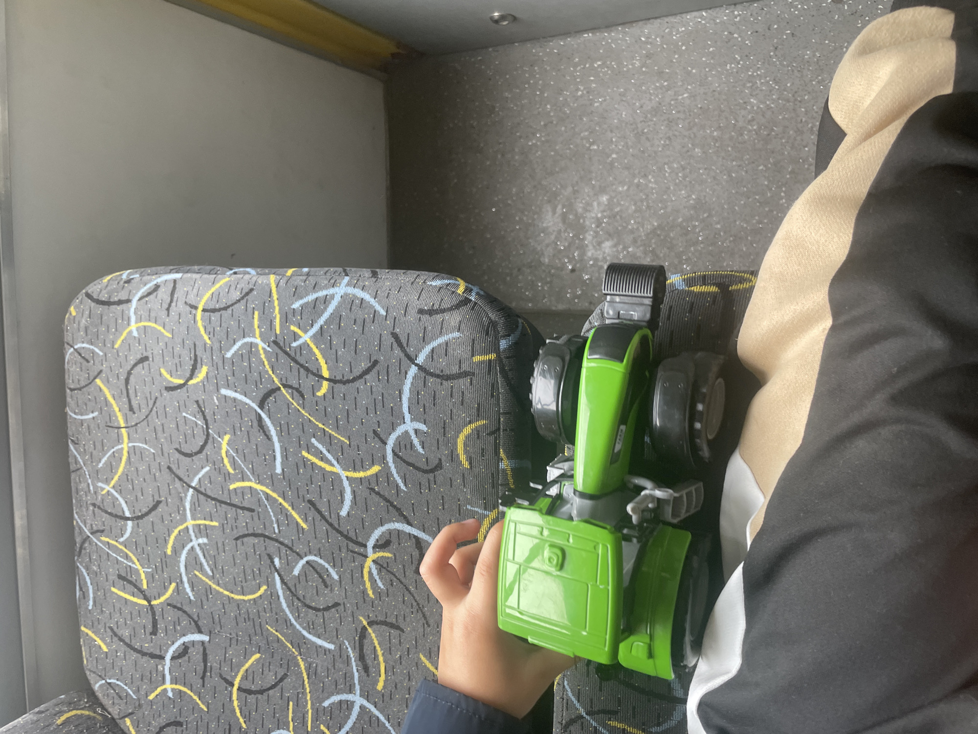 A child's hand is visible, holding a green toy tractor on a seat with a grey and yellow patterned fabric. The tractor is partially obscured by the child's clothing, which is black and tan. The background shows a textured floor and a yellow stripe on the right side.