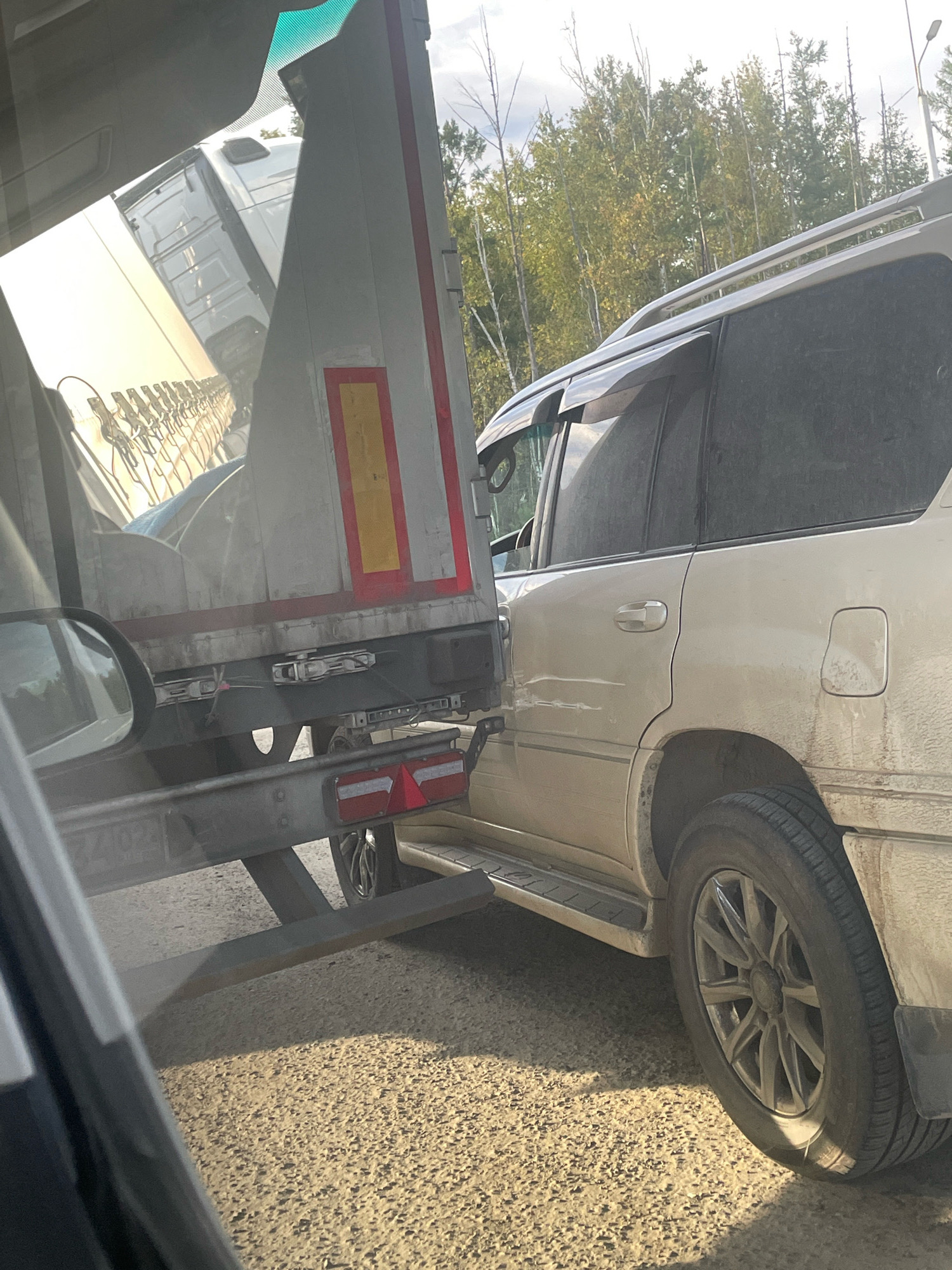 A beige SUV, possibly a Toyota Land Cruiser, is positioned at a right angle to the trailer of a large truck. The rear passenger side of the SUV is damaged, with visible creasing and deformation in the body panel extending from the rear door to the wheel well. The SUV's rear wheel is turned slightly outward. The trailer of the truck, a white and silver semi-trailer, appears to have sustained some damage on its rear underside, including a bent metal bracket. The trailer also has reflective red and yellow markings. The scene is set on a gravel road, likely in a rural or semi-rural area, with a line of trees, some showing autumn colors, in the background. The sky is overcast with patches of sunlight. The image appears to be taken from inside another vehicle, with a portion of a side mirror and windshield visible in the foreground. The license plate of the truck's trailer is partially visible, showing the numbers "7402". The setting is identified as Sivaki, Russia.