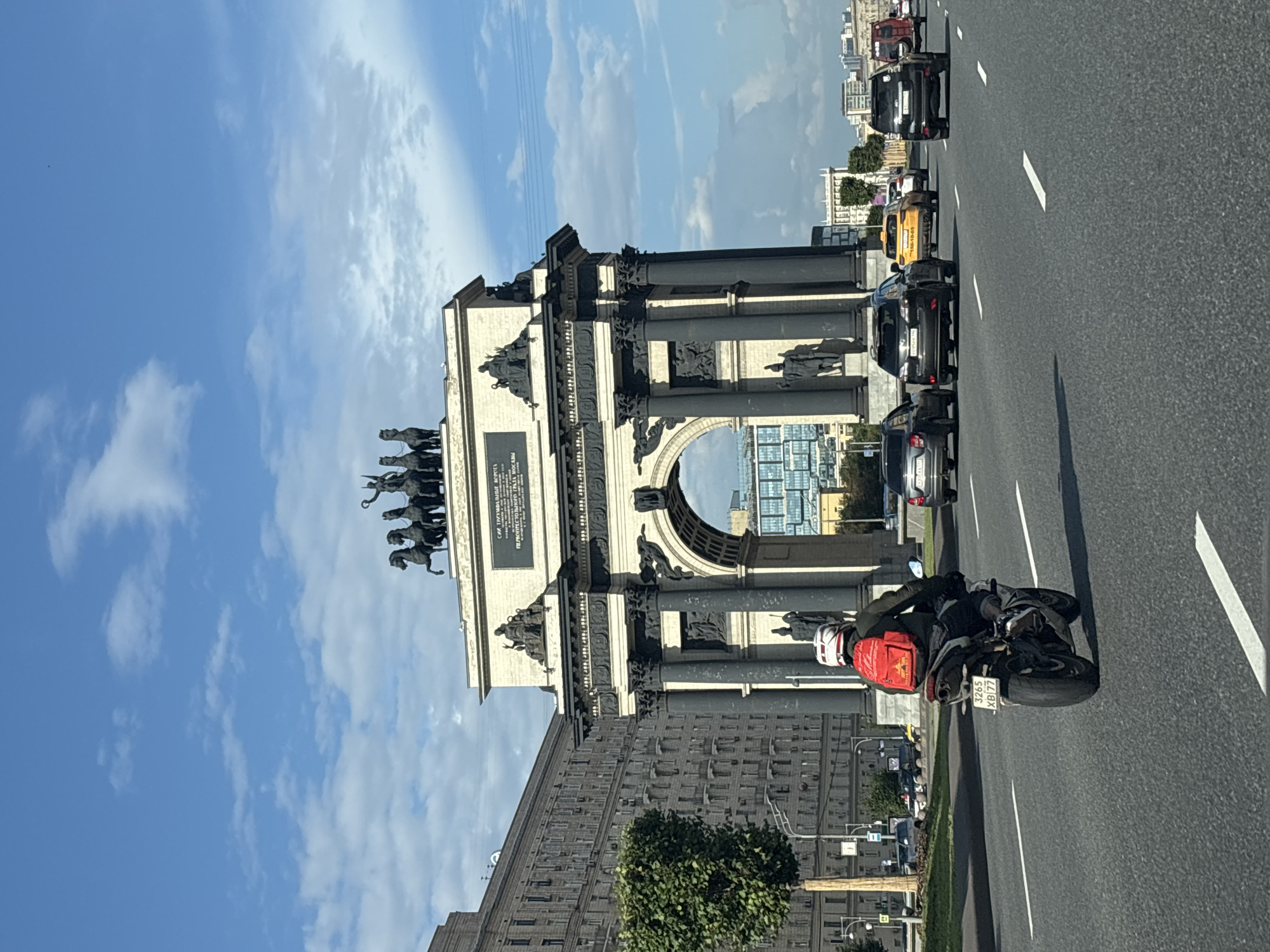 The image captures a frontal view of the Triumphal Arch (Triumfal'naya Arka) in Moscow, Russia, on a clear day with scattered clouds. The arch is a monumental structure, adorned with sculptures and inscriptions, with a quadriga crowning its apex. In the foreground, a person rides a motorcycle with a red backpack. The motorcycle's license plate is visible, reading "3265 XB 77." Several cars are driving on the road in front of the arch, including a yellow taxi. To the left of the arch, a large, multi-story building with many windows stands prominently. A small, well-trimmed tree is situated between the building and the road. The scene depicts a busy urban environment with architectural landmarks and active traffic.