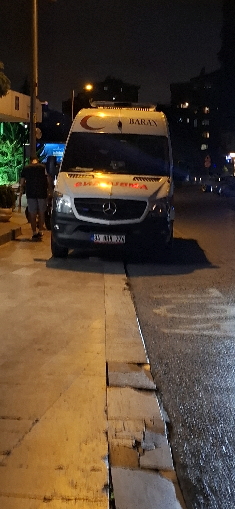 An ambulance, identified as "BARAN", is parked on a street at night in Kadıköy, Turkey. The ambulance, a white Mercedes-Benz Sprinter, has its hazard lights illuminated and a red crescent logo on its side. A man in a dark t-shirt and shorts is standing near the ambulance, facing away from the camera. The street appears to be wet, suggesting recent rain or a damp surface. The scene is illuminated by streetlights and the ambulance's own lights, casting reflections on the pavement. The surrounding environment shows buildings in the background, typical of an urban setting. The license plate of the ambulance reads "34 URN 774".