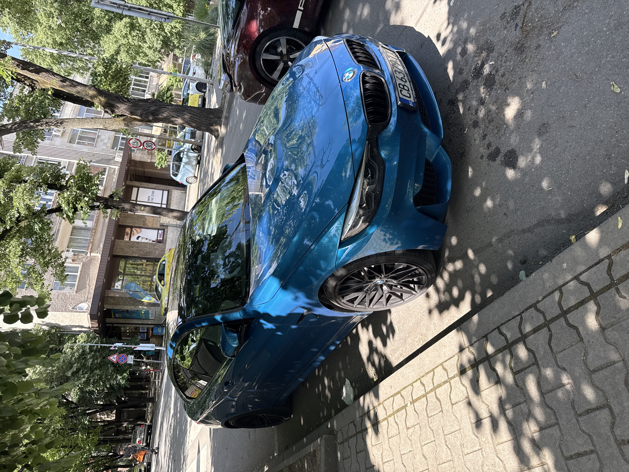 The image captures a bright, sunny day on a city street in Stara Zagora, Bulgaria. The central focus is a blue BMW parked along the sidewalk. The car's license plate reads "CB 6379 TK". A burgundy-colored vehicle is partially visible beside it. The sidewalk is paved with interlocking concrete blocks. The street is bordered by trees, casting dappled shadows on the pavement and the cars. In the background, buildings are visible, including one with a storefront featuring the word "FLO". Street signs and traffic signals are also present, indicating an urban environment. A few other vehicles are visible further down the street. Pedestrians are also seen further down the street.