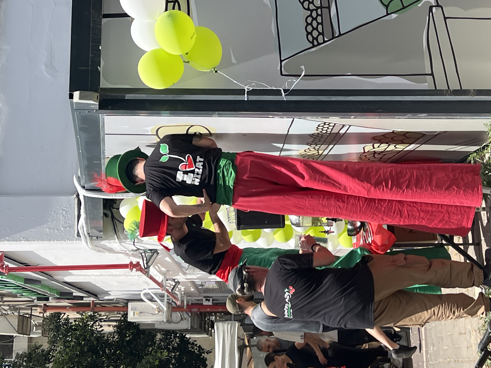 The image captures an outdoor event on a bright, sunny day, likely in an urban environment. Several people, mostly men, are present, suggesting a public gathering or festival.
In the center, a man stands prominently, wearing distinctive attire: bright red, possibly stilt-like or oversized pants, a green sash, a black t-shirt with a cherry-like logo and the word "NIZAT" in white text, and a green hat adorned with a red feather. He appears to be looking down at another man, also wearing a green hat and a red sash/belt, who is partially obscured.
Other individuals are visible in the foreground and background. One man in the lower right foreground wears a black t-shirt with a similar cherry-like logo and the word "REVIVAL" below it, paired with khaki pants. Another person in a grey t-shirt is partially visible behind him. Further back, another man in a black t-shirt and dark pants is also present.
The scene features festive elements such as clusters of white and lime green balloons. What appear to be market stalls or tables, some draped with red or green fabric, are also visible. The background includes a white building facade with black and white line art decorations, as well as red industrial-style pipes and some greenery from trees or bushes, indicating an outdoor setting within a built-up area. The general atmosphere suggests a casual, lively community event or market.