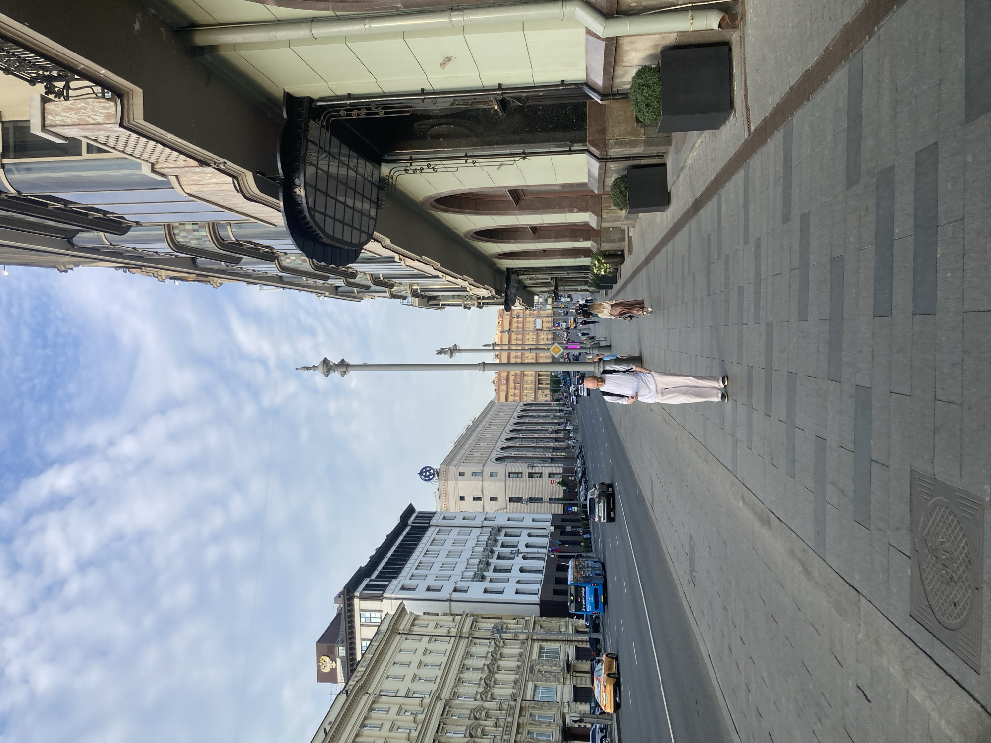 The image depicts a street scene in Moscow, Russia, likely on Tverskaya Street, a major thoroughfare known for its grand architecture. The perspective is from a sidewalk looking down the street. On the right side of the frame, a light green building with a decorative entryway and arched windows dominates the foreground. Large black planters with green shrubs line the sidewalk in front of this building. Further down the street, on the left, is a row of multi-story buildings with ornate facades, typical of Moscow's historic center. One building features a prominent double-headed eagle emblem, suggesting it might house a government institution or a historic landmark.
A paved sidewalk occupies the lower half of the image, with a street running alongside it. Several cars are visible on the street, including a yellow taxi and a blue bus with Russian text. A man in white pants and a white shirt is walking on the sidewalk away from the camera, holding a phone. Further down the sidewalk, a woman in a long skirt and a light-colored top is also walking away from the camera. The sky is partly cloudy with a soft, diffused light, indicating it might be morning or late afternoon. The overall impression is a bustling urban environment with a blend of historic and modern elements.