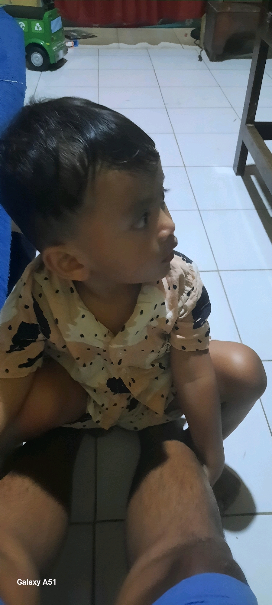 The image shows a young child, likely a toddler, sitting on a tiled floor. The child is wearing a light-colored shirt with a pattern of black dots and abstract shapes. The child is looking off to the right side of the frame. The floor appears to be indoors, with white square tiles and visible grout lines. In the background, there are glimpses of furniture and toys, including a green toy truck. The lighting suggests an indoor environment, possibly a living area or playroom. The bottom left corner of the image displays "Galaxy A51". This image was taken in Jatitengah, Indonesia.