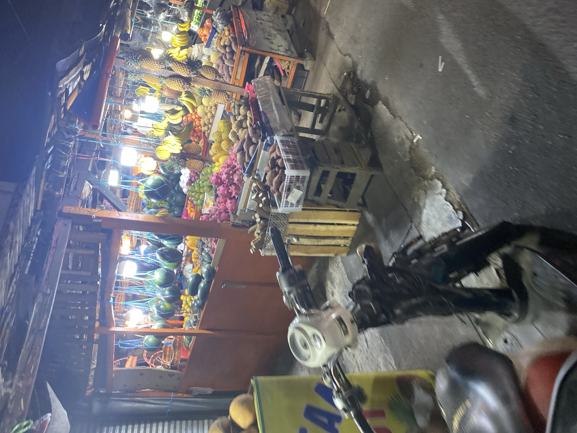 The image depicts an outdoor fruit stall at night in Tangerang, Indonesia. The stall is brightly lit with artificial lights, illuminating a wide variety of fruits. Watermelons are hung from the ceiling, alongside bunches of bananas and pineapples displayed vertically. Other fruits visible include dragon fruit, papayas, and what appear to be sweet potatoes or yams in baskets. The stall is constructed with wooden frames and covered with a corrugated metal roof. In the foreground, the handlebars and front portion of a white scooter are visible, suggesting a mode of transportation that has been parked nearby. The ground is paved with asphalt and shows signs of wetness. The overall impression is a bustling night market scene, common in Indonesian cities.
