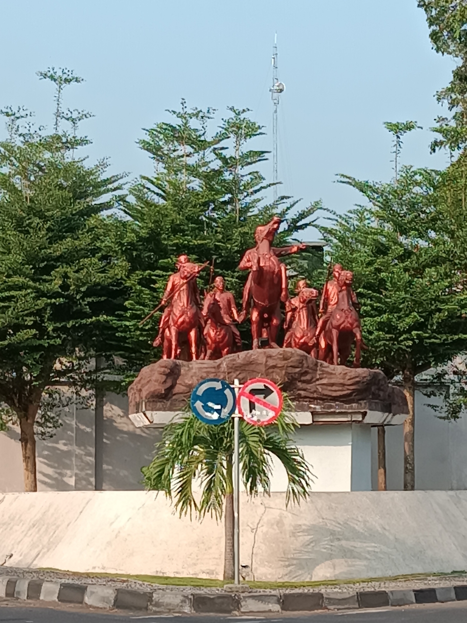 In Imogiri, Indonesia, a bronze equestrian statue of five figures stands atop a rocky pedestal in a public area. The central figure, pointing with his right arm, is positioned on a horse with its head raised. To his left and right, four other figures on horseback are also depicted in various stances. The sculpture is surrounded by lush green trees, and a tall communication tower is visible in the background against a clear blue sky. In the foreground, two traffic signs are mounted on a pole: one circular blue sign with three arrows indicating a roundabout, and a red circular sign with a diagonal bar across a right-pointing arrow, signifying no right turn. A palm tree with fronds arches over the signs. The base of the pedestal is a textured, reddish-brown surface, and the entire monument is situated on a raised concrete platform. The scene appears to be a roadside monument or a park setting.