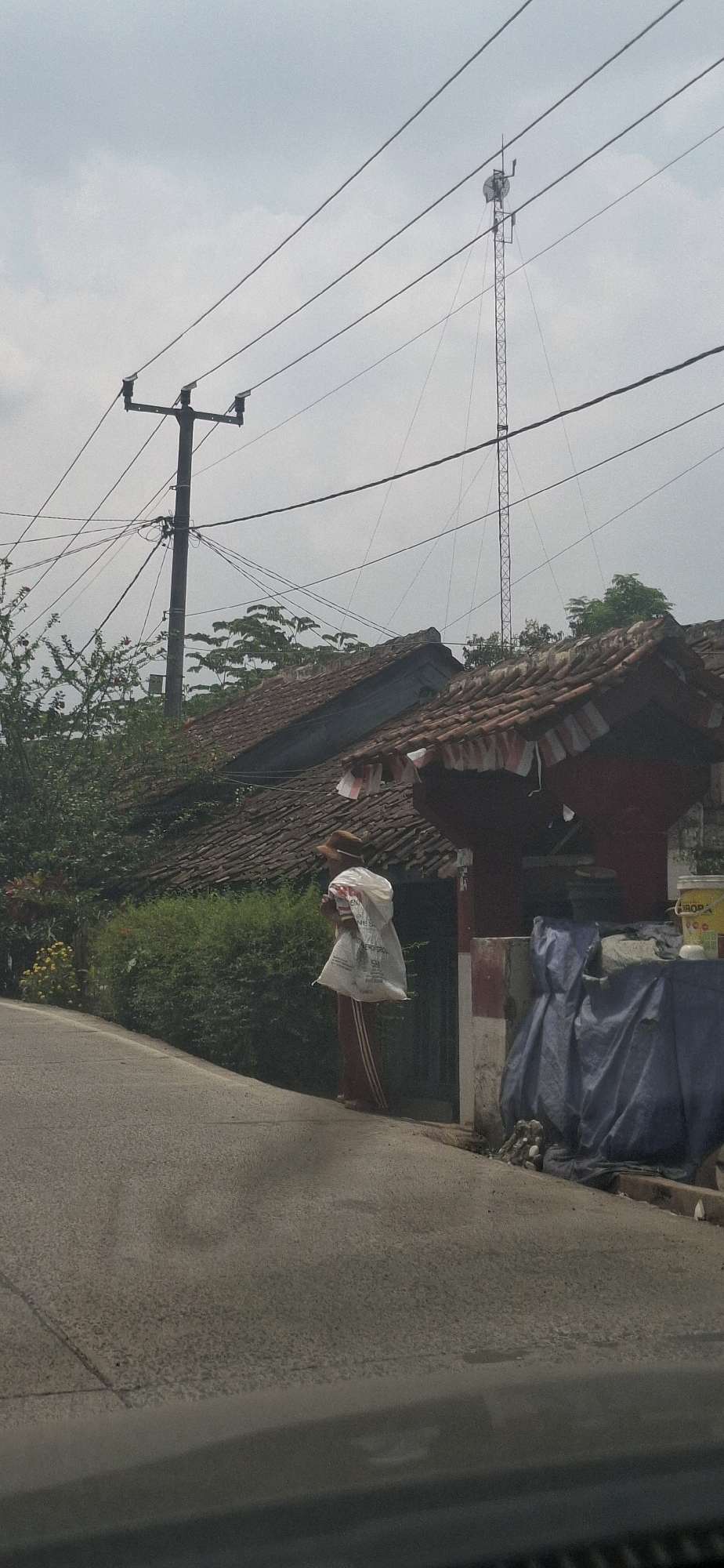 The image depicts a street scene in Cibinong Hilir, Indonesia, under an overcast sky. In the foreground, a paved road curves slightly to the left. A person, wearing a hat and a red and white striped garment, walks along the side of the road carrying a large white plastic bag over their shoulder. They are positioned near a building with a tiled roof, adorned with small Indonesian flags. Adjacent to the building, a blue tarp covers various objects. Behind the building, lush green foliage and trees are visible. In the middle ground, a utility pole with multiple electrical wires stretches across the frame, and further in the distance, a tall transmission tower with antennas stands against the sky. The scene suggests a residential or semi-rural area with visible infrastructure.