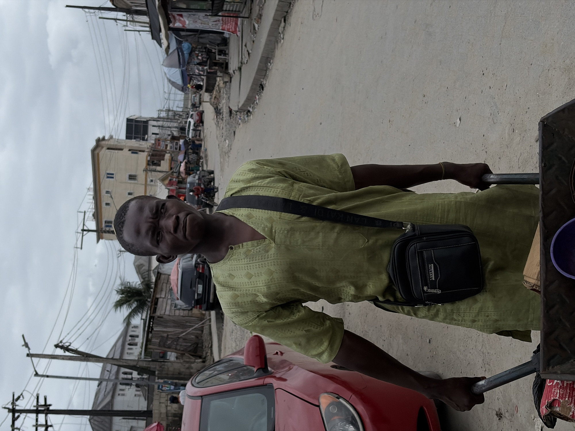 In Igboefon, Nigeria, a Black man walks down a dusty, debris-strewn street pushing a cart. He is wearing a light green tunic with a v-neck and an ornate diamond pattern. A black crossbody bag is slung over his shoulder. His left hand grips the cart's handle. The cart contains various items, including what appears to be cardboard and a purple bowl. To the left of the man, a red car is parked. The background shows buildings, utility poles with numerous wires, market stalls with umbrellas, and other vehicles. The sky is overcast.