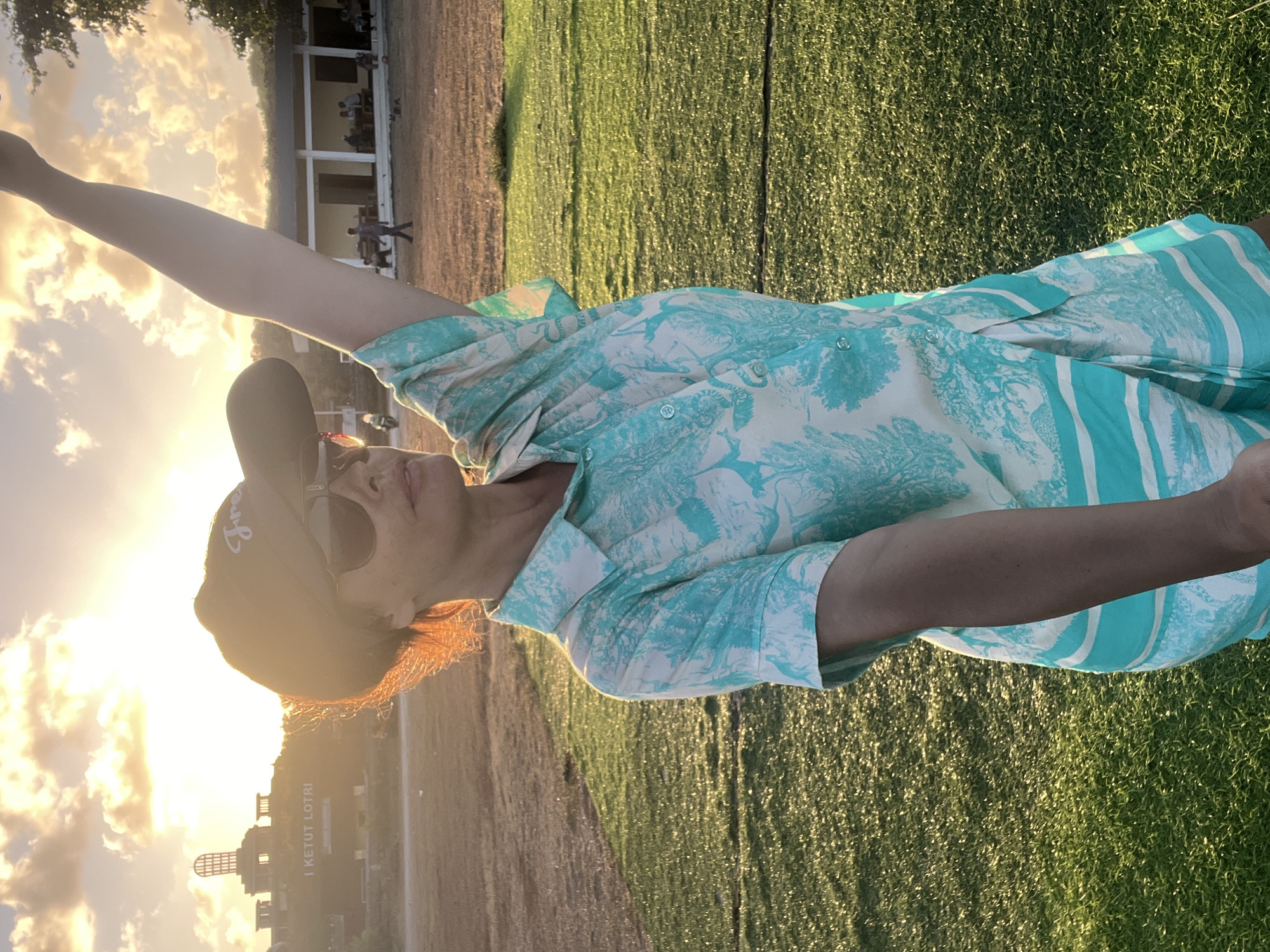 A woman stands on a grassy field, facing right, with her left arm raised high. She wears a black baseball cap, sunglasses, and a short-sleeved button-down shirt with a white and turquoise pattern. The sun is setting, casting a warm glow and long shadows across the scene. In the background, a building with a distinctive tower structure and text that reads "I KETUT LOTRI" is visible on a hill. Other people and buildings are also present in the distance, and a motorcycle is parked to the left. The setting appears to be a public space in Kuta Selatan, Indonesia.