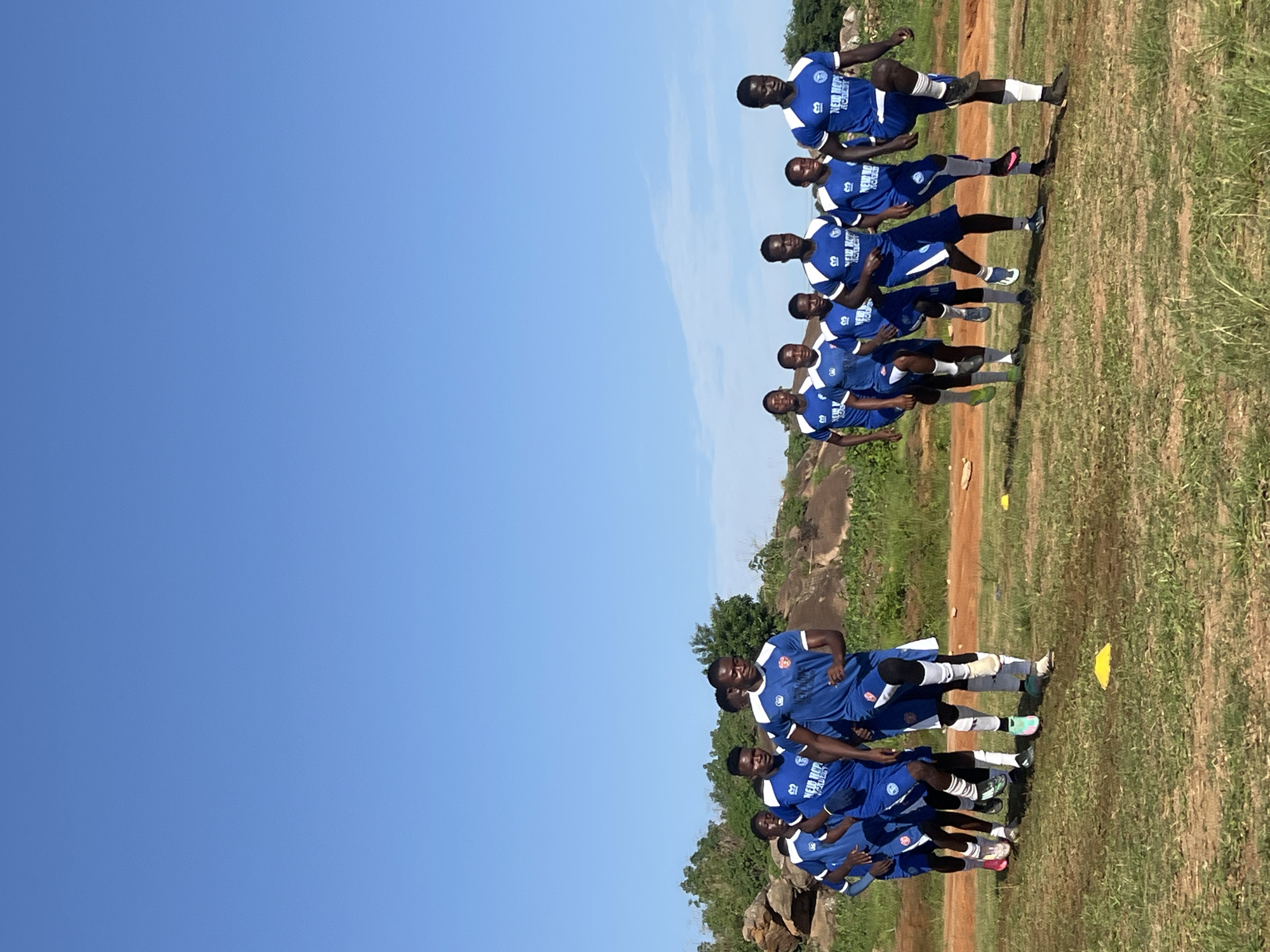 A group of young male soccer players, all wearing blue and white uniforms with "NEW HOPE ACADEMY" inscribed on them, are captured in motion outdoors. They are in the process of a high-knees drill, with their legs raised in unison. The players are arranged in a staggered line, moving from left to right across the frame, with the focus on the front row. The environment suggests an outdoor training facility, possibly in a semi-arid region, characterized by sparse, dry grass, exposed reddish-brown earth, and scattered rock formations in the background. The sky above is a clear, bright blue, indicating sunny weather. The scene is consistent with a training session taking place at a soccer academy in Jos, Nigeria.