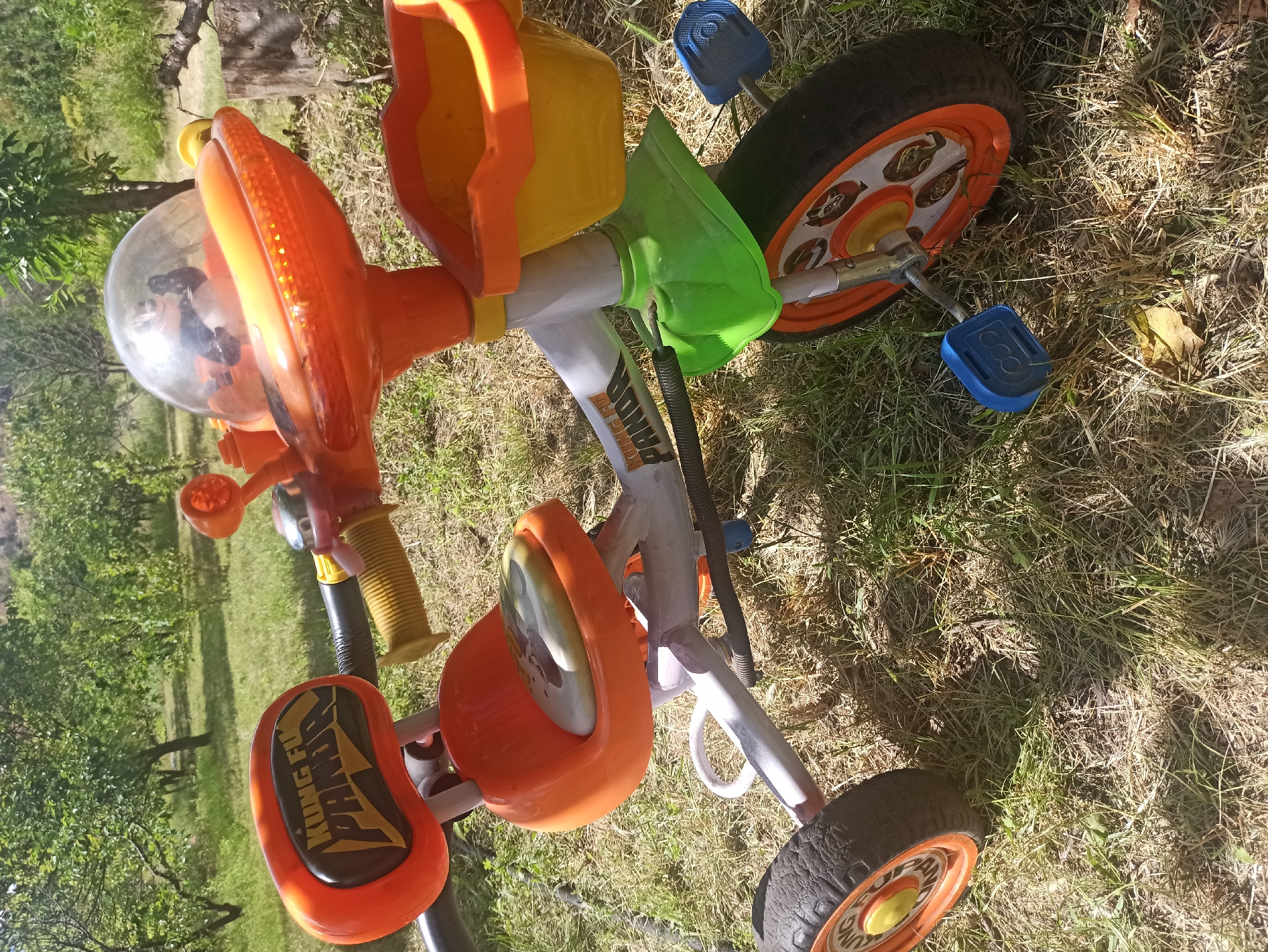 This media file shows a close-up, overhead view of a child's tricycle lying on its side in a grassy outdoor setting. The tricycle is brightly colored, with prominent orange, yellow, and green plastic components. Key visible parts include the handlebars, a steering wheel covered with a clear plastic dome containing some black objects (possibly toys), a seat with "KUNG FU PANDA" text in yellow and black, pedals, and wheels. The rear wheel is partially visible.
The tricycle appears to be an object, possibly discarded or temporarily placed on the ground, rather than actively being used by a child. The background consists of green grass and some dry leaves, with hints of trees and foliage suggesting a natural, possibly park-like or backyard environment. The lighting indicates it's daytime, with sunlight casting shadows, suggesting a sunny day. There are no people visible in the frame. The visible text on the seat reads "KUNG FU PANDA". The overall impression is one of a bright, colorful toy in a slightly unkempt outdoor space.