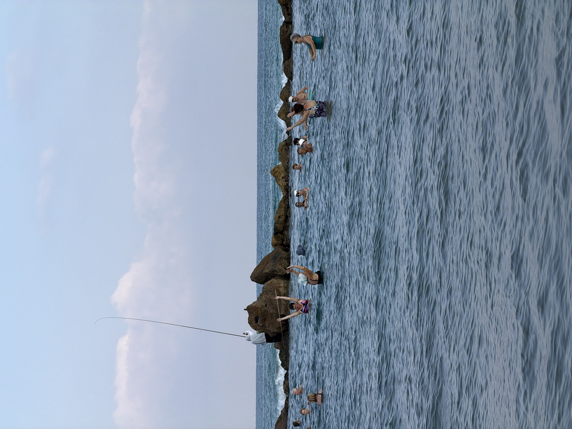 This is a photograph taken at the Bat Yam beach in Israel, featuring a group of people in the ocean, engaged in what appears to be a water aerobics or exercise class. The water is calm with gentle ripples. In the background, rocky structures jut out of the sea, with a person standing on one of them holding a fishing rod. Several individuals, mostly adults, are visible in the water, with some actively participating in the exercises, raising their arms. The sky is partly cloudy with a pale blue hue. The overall scene depicts a recreational activity taking place in a coastal environment.