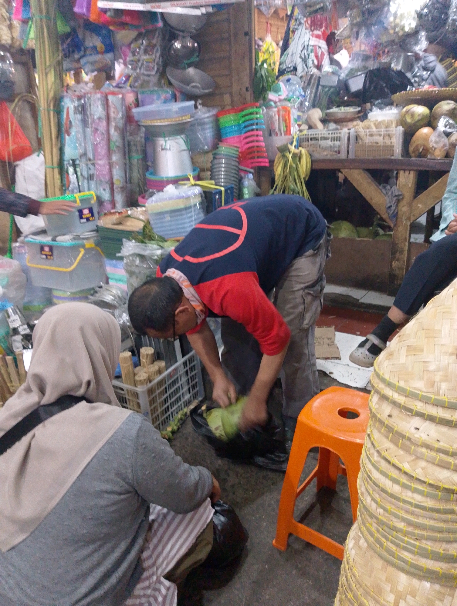 The image depicts a bustling market scene in Lembang, Indonesia, likely a local produce or household goods market. In the foreground, a person wearing a beige hijab and a grey sweater sits with their back to the camera. They are partially obscured by a pile of woven baskets stacked to their right. To the left, a person's hand, likely belonging to someone off-camera, reaches out towards a set of plastic containers.
The central focus is a man in a blue and red shirt and grey pants, bending over to handle what appears to be a green vegetable, possibly a melon or squash. He is surrounded by various market goods, including more plastic containers, woven baskets, and what look like bundles of sticks. Behind him, a wooden stall displays an assortment of goods, including fruit (coconuts and other round fruits), and stacked items wrapped in plastic. The overall environment suggests a lively, somewhat cluttered, and vibrant marketplace. The ground appears to be concrete, typical of such venues. The lighting suggests it is daytime, with some shadows indicating overhead illumination or light filtering from an unseen source.