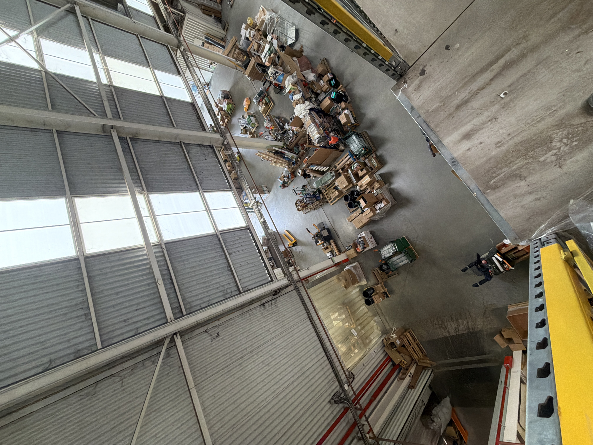 The image depicts an aerial view inside a large industrial building in Stroevo, Bulgaria. The building has a high ceiling with corrugated metal siding and a series of windows. The floor is concrete and appears to be cluttered with various objects, including boxes, pallets, and other materials indicative of warehouse storage. A person is visible on the floor, dressed in dark clothing. Shelving units are visible along the right side of the image.