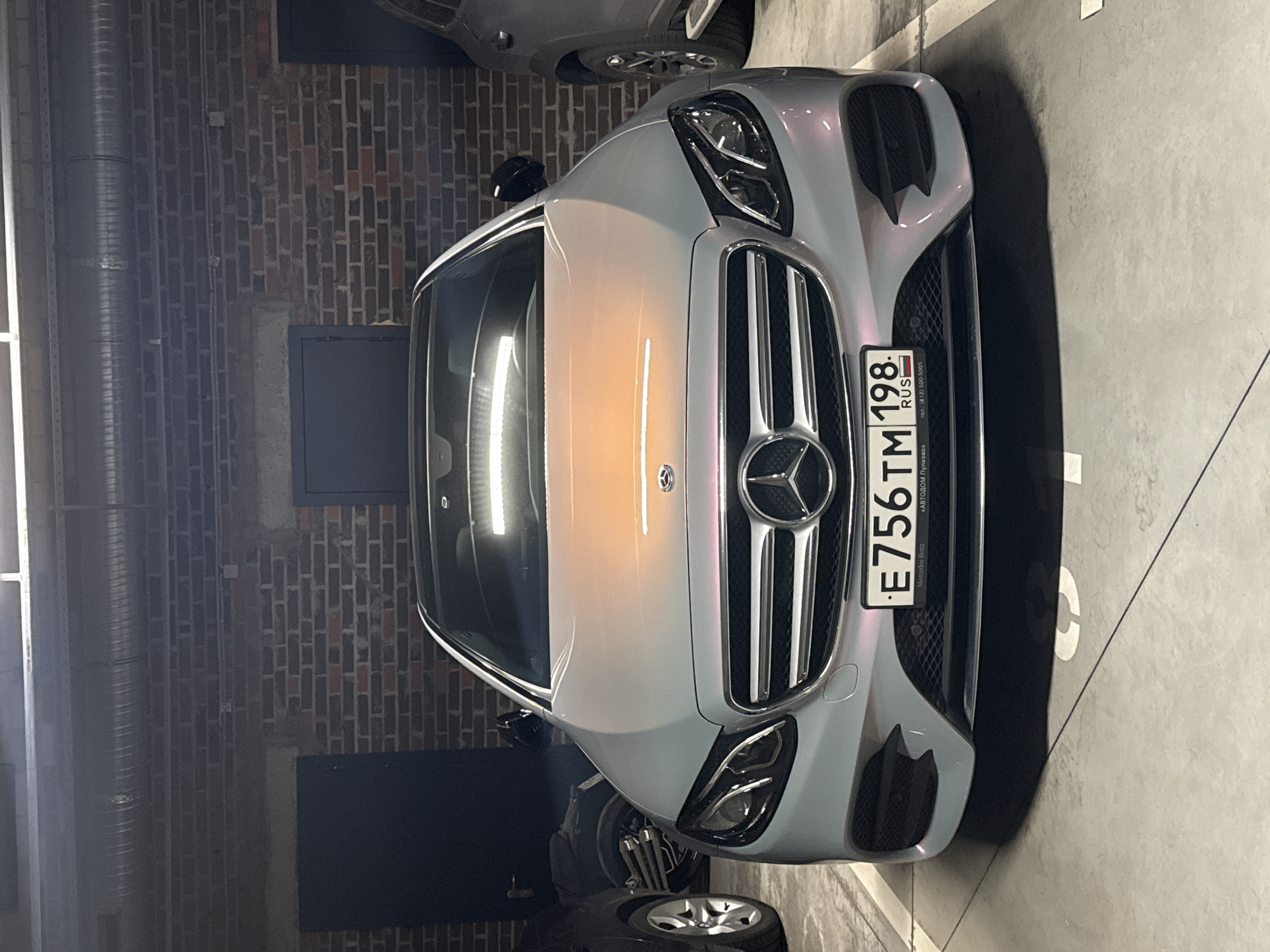 An eye-level shot captures the front of a light gray Mercedes-Benz car, likely an E-Class, parked indoors. The car's surface exhibits an iridescent quality, reflecting hues of silver, light blue, purple, and orange-pink from overhead lights. The prominent Mercedes-Benz emblem is visible on the grille.
The car displays a Russian license plate, "E756 TM 198 RUS," with "Mercedes-Benz" printed below it. To the right of the plate, text reads "«АВТОДОМ Пулково»" and "тел.: (812) 500-5005," indicating a Mercedes-Benz dealership located in Pulkovo, Saint Petersburg, Russia.
The setting appears to be a parking garage or basement, characterized by a dark brick wall on the left side of the frame and a concrete floor with white markings where the car is parked. A large, corrugated metallic pipe runs vertically along the brick wall. The front tire and part of the bumper of another vehicle are partially visible at the top and bottom edges of the image, suggesting close parking. Artificial lighting illuminates the scene, casting reflections on the car's paintwork and windscreen. No people are visible, and the scene depicts a static, parked vehicle.
