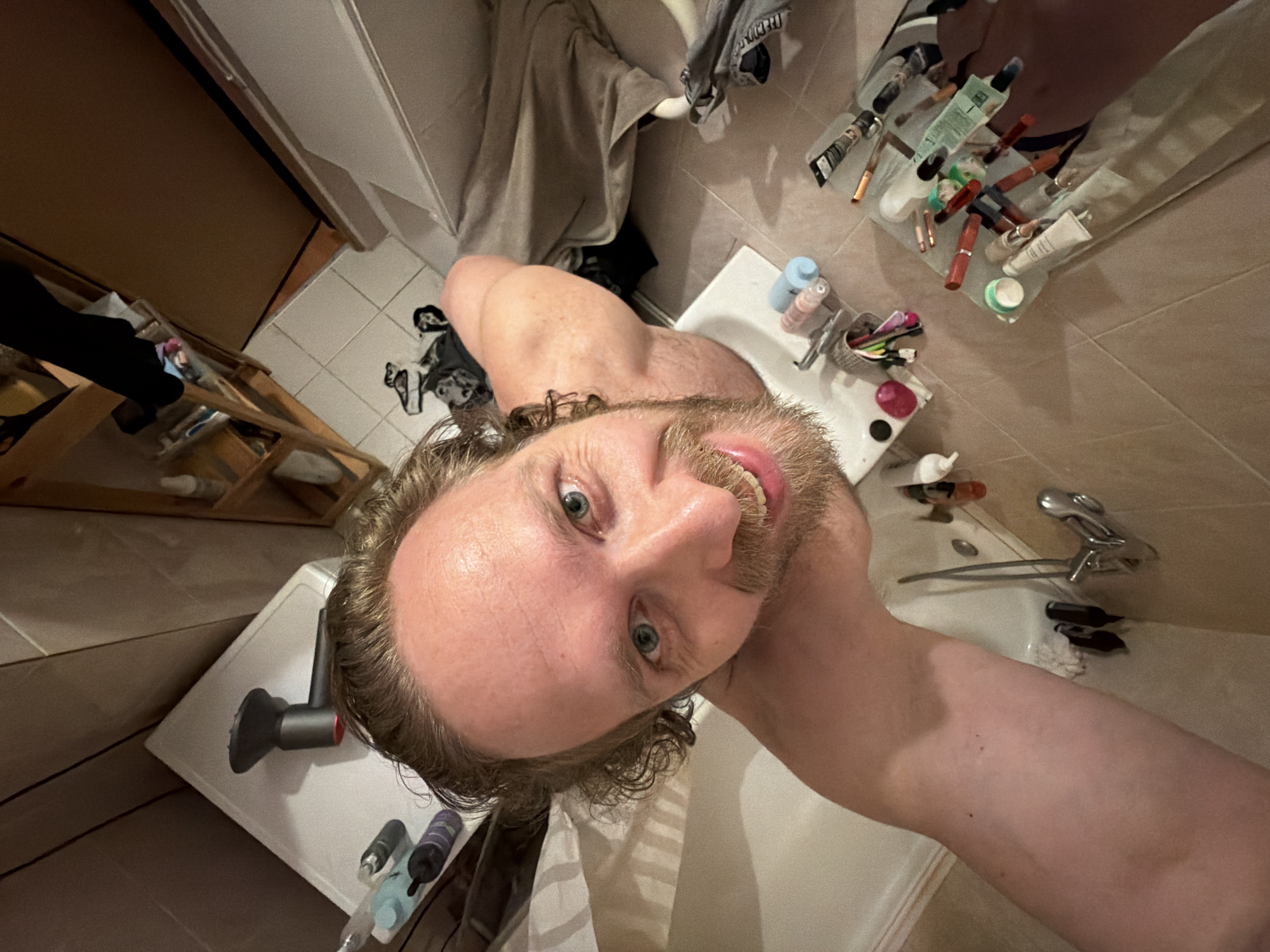 A man is positioned in a bathroom, looking upwards into the camera, with his head tilted. He has fair skin, light brown hair, and a short beard. His mouth is slightly open, revealing his teeth. The bathroom features a tiled floor and walls. There is a sink with a faucet and various toiletries, including tubes of creams and makeup items, are scattered around the sink area. A hairdryer rests on a counter, and there's a wooden shelf with items on it. The location is likely a private bathroom and cannot be confirmed to be in Saint Petersburg, Russia based on the image alone.