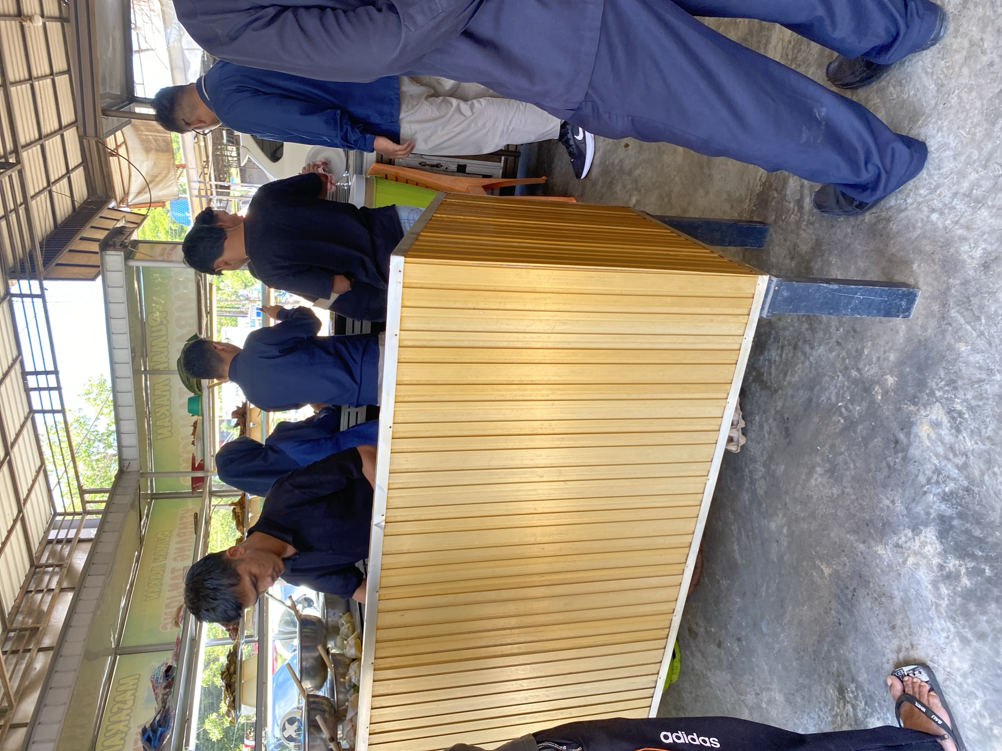 The image depicts a food stall in Banda Aceh, Indonesia, likely an outdoor or semi-outdoor establishment given the visible roof structure and natural light. The central focus is a food counter made of light-colored wooden slats, with a metal trim along the top. Behind the counter, several individuals are visible.
In the foreground, a young man wearing a black polo shirt is looking down, seemingly preparing food or serving customers. To his right, a person wearing a dark blue hijab and a dark top is also positioned behind the counter. Further to the right, another individual in a black t-shirt is visible. The background reveals more people, including one in a blue uniform and beige pants, wearing glasses. Another person, partially visible on the far right, is wearing a dark blue uniform and dark pants.
On the shelves behind the counter, various food items are displayed in containers and trays. Some metal bowls are also present on the counter. The environment appears to be a busy food service area, with evidence of ongoing operations. The floor is concrete. Signs with text in Indonesian are visible on the wall behind the counter, suggesting this is a commercial establishment. The overall scene captures a moment in the daily life of a food vendor in Banda Aceh.