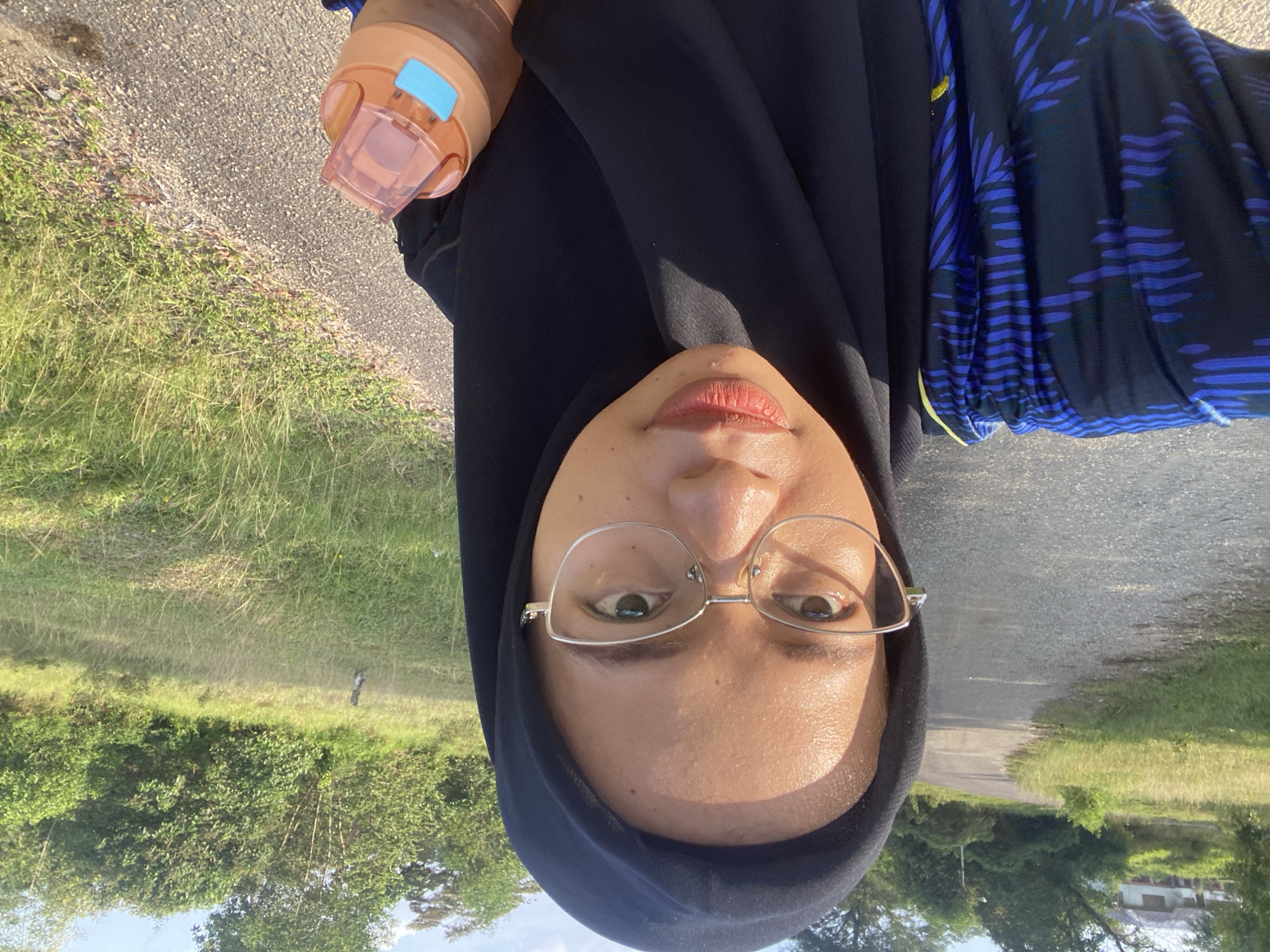 A woman wearing a black hijab and glasses is taking a selfie on a paved road lined with dry grass and trees. She is wearing a dark blue garment with a pattern of blue and black lines and leaf-like shapes. In her right hand, she holds an orange water bottle with a blue lid. The road curves to the left in the background, leading towards a distant building and more trees. The sky is partly cloudy and the sun appears to be shining. The location is identified as Gudang Batu, Indonesia.