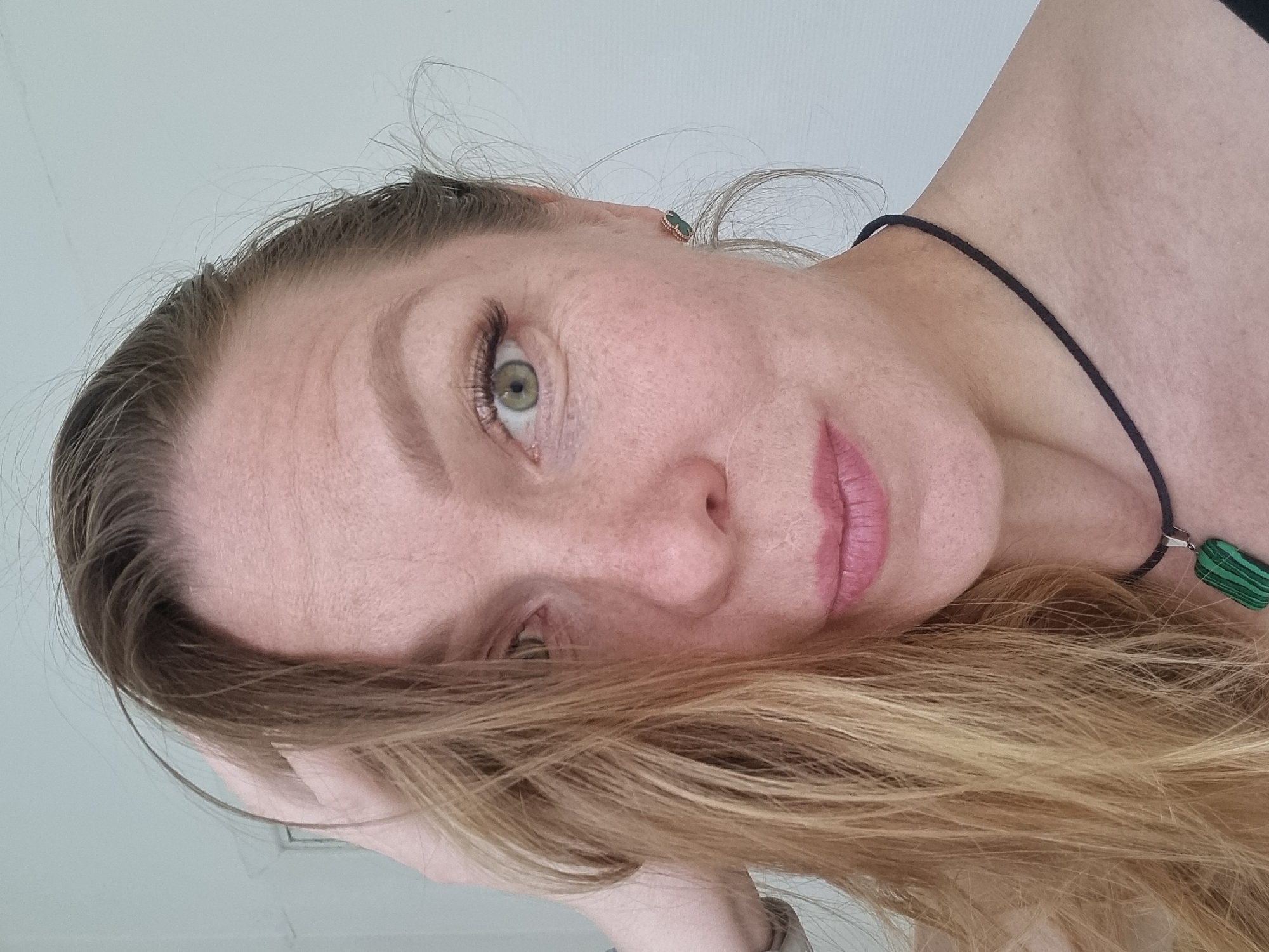 A close-up, angled portrait shows a woman with light skin, blonde hair, and green eyes. She has visible freckles on her face. She's wearing a black cord necklace with a green pendant and green earrings. The background appears to be a plain white or light-colored wall. There is no evidence to suggest the location is in Kemul, Russia.