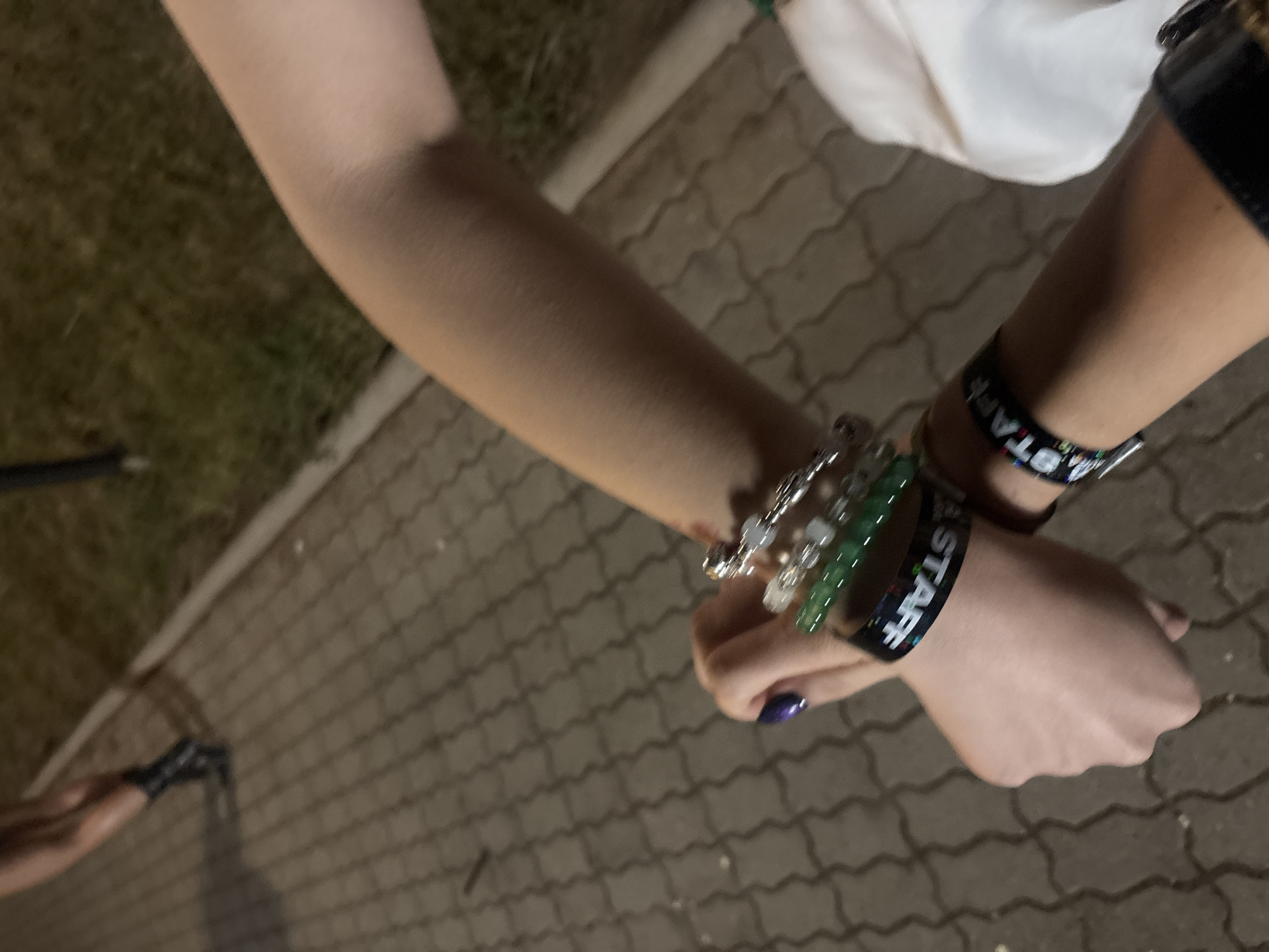 The image shows a close-up of two arms wearing bracelets and wristbands. The background is a paved walkway and grassy area, suggesting an outdoor urban environment. One wristband clearly displays the word "STAFF" in white lettering on a black background. The other arm has a stack of bracelets, including some with green beads and others with clear beads, as well as a black wristband with "STAFF" visible. The presence of "STAFF" wristbands indicates the individuals are likely working at an event or venue. The overall lighting suggests it is evening or night.