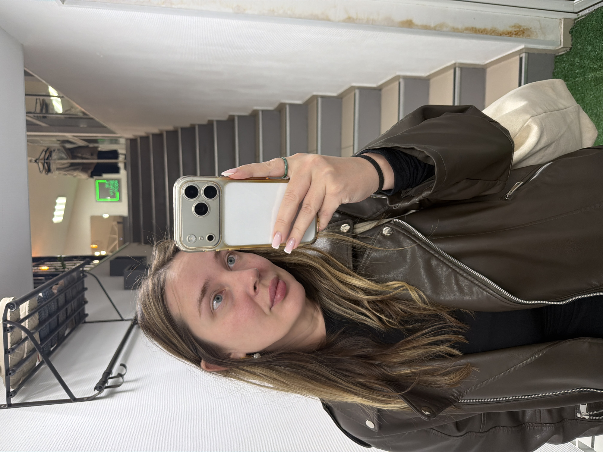 A woman takes a selfie in front of a mirror. The mirror reflects a staircase, a white bag, and part of a clothes rack. The woman wears a brown leather jacket, a black shirt, and a ring on her left hand. She holds a smartphone in her right hand. The location may be a shop or store. This image may have been taken in the city of Chaikovsky, Russia.