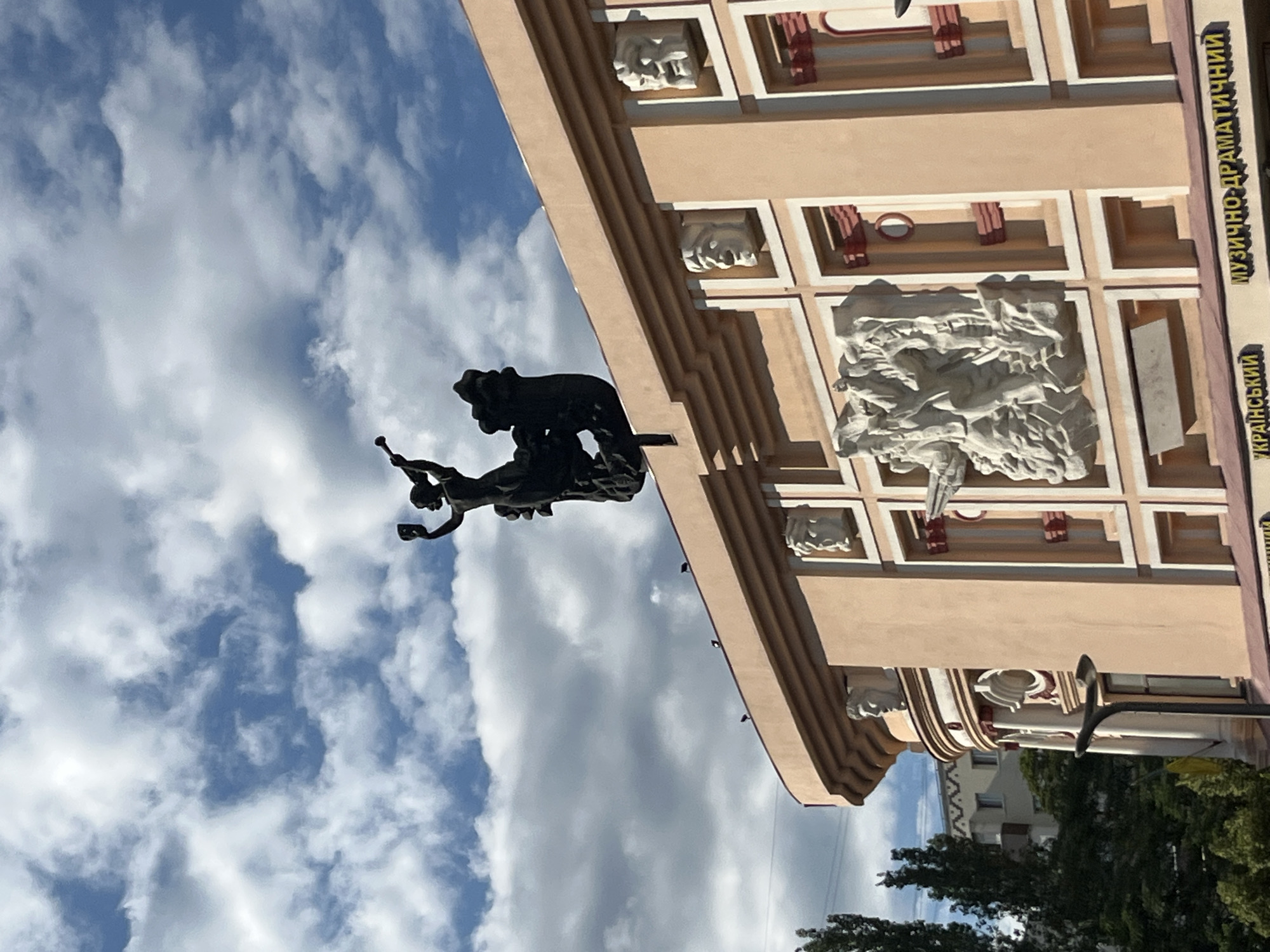 The media file captures a view of a building's facade against a cloudy sky. A dark, metal sculpture of what appears to be two figures wrestling or in a struggle is prominently featured against the sky. The building itself is light-colored with decorative architectural elements, including what look like bas-relief sculptures of faces and intricate patterns. The facade has a row of square recesses with red accents. Text is visible vertically along the right side of the building. The inscription reads "МУЗИЧНО-ДРАМАТИЧНИЙ" and below it, "УКРАЇНСЬКИЙ". The lighting suggests it is daytime, and the presence of clouds indicates overcast or partly cloudy weather. The image is taken from a low angle, looking upwards towards the building and sky. The overall scene conveys a sense of grandeur and artistry associated with the architecture and sculpture. The location is identified as Dnipro, Ukraine.