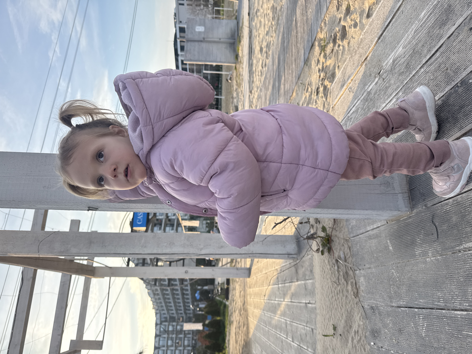 A young child with blonde hair in pigtails, wearing a pink hooded puffer jacket, pink pants, and pink shoes, is climbing on a wooden structure, likely a fence or railing. The structure is painted white. The child appears to be gripping the horizontal beam of the structure, and her body is parallel to the ground. The setting is outdoors, possibly at a beach or boardwalk, with wooden planks visible on the ground. In the background, there are buildings, and the sky is partially visible, suggesting it's daytime. The image appears to be taken in Kranevo, Bulgaria, based on the surrounding buildings' architecture and layout.