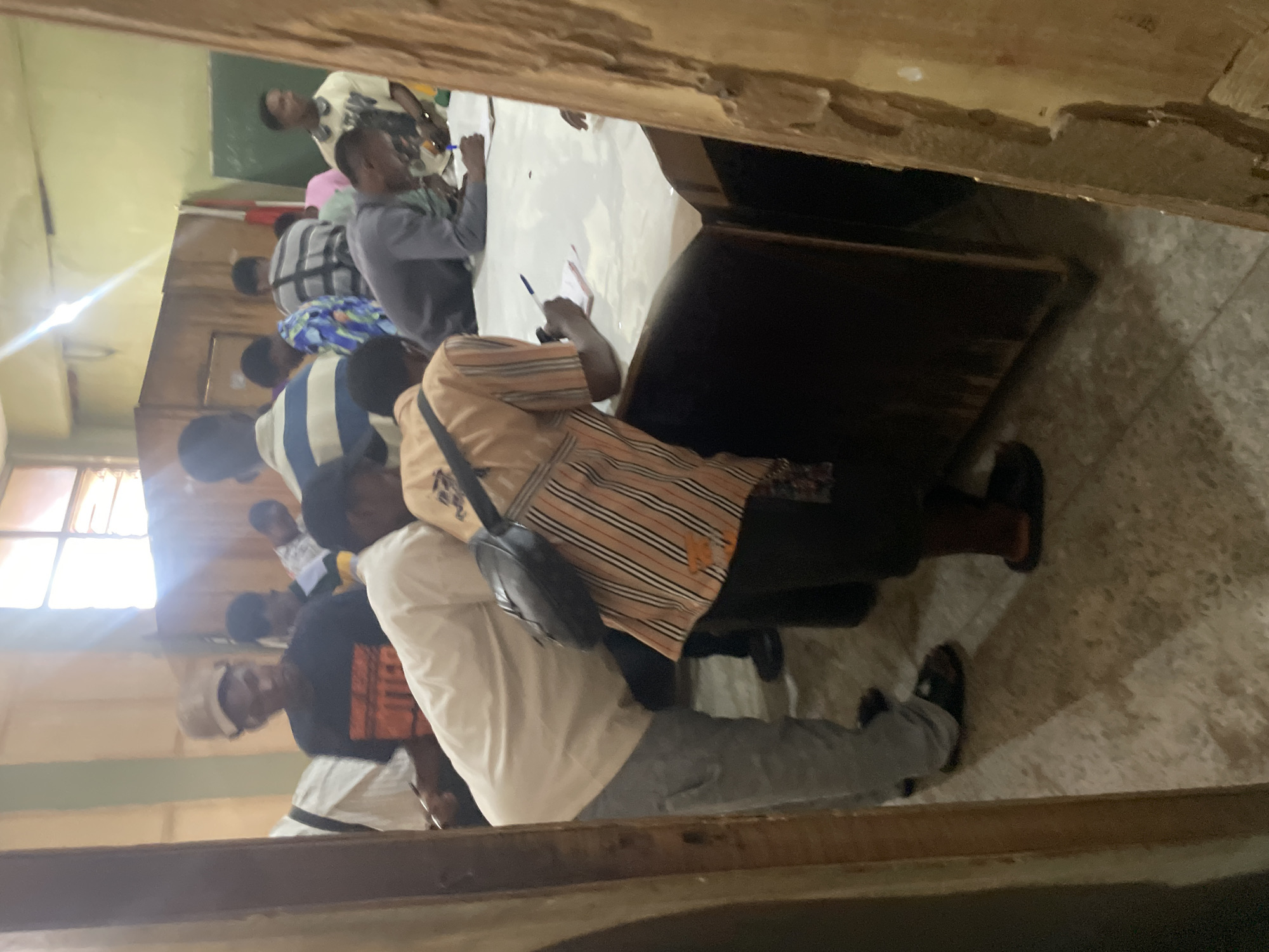 The image depicts an indoor scene, likely a classroom or meeting hall, with a group of young Black men gathered around a large, light-colored table. The room appears to be in a state of disrepair, with peeling paint on the walls and a rough wooden door frame in the foreground. Sunlight streams in from a window with multiple panes, casting glare on the wall and floor.
Several individuals are actively engaged in writing on papers spread across the table. One person in the foreground, wearing a light-colored long-sleeved shirt and gray pants, is bending over the table, holding a pen and appearing to write. To their left, another individual in a black t-shirt with an orange design and a tan baseball cap is also leaning over the table, though their action is less clear. Another person further back, wearing a striped shirt, is also writing.
Other individuals are standing or leaning in, observing or waiting. The overall atmosphere suggests an organized activity, possibly an examination, registration, or a meeting where documentation is being handled. The floor is tiled with a speckled pattern. The presence of a blackboard or similar surface on the wall in the background indicates an educational or official setting. The visual cues suggest the location is within Ihiagwa, Nigeria, likely a public institution such as a school or community center.