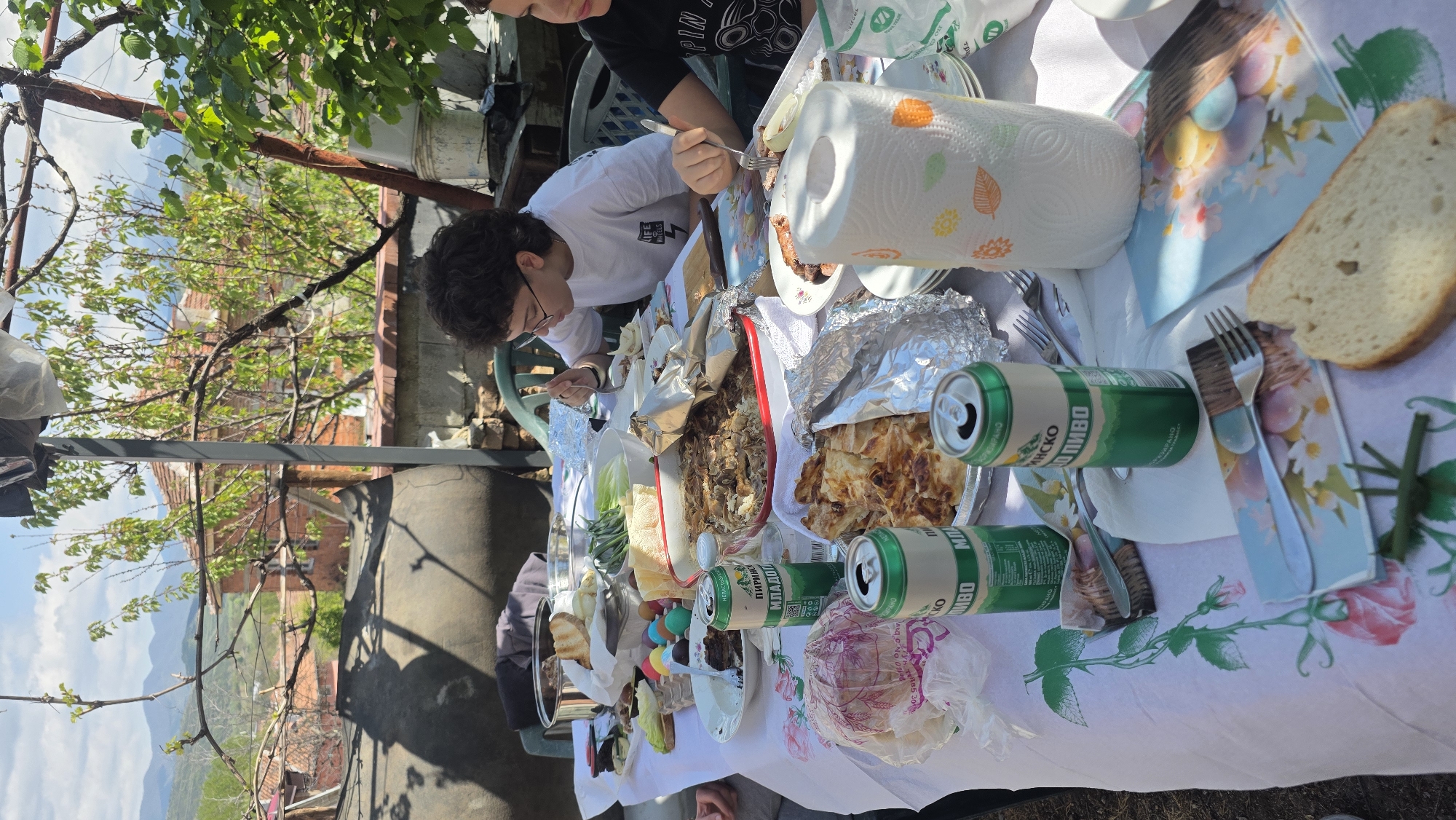 The image depicts an outdoor gathering, possibly a meal, taking place in Katuntsi, Bulgaria. Two people are visible at the table: one wearing glasses and a white t-shirt is actively eating with a fork. The other person is wearing a dark shirt and is partially visible.
The table is covered with a white tablecloth adorned with floral patterns and Easter egg motifs. Various food items are present including meat in foil, pastry, slices of bread, salads and cakes. There are also numerous cans of пиво (beer) visible on the table.
The setting seems to be an outdoor patio or garden with trees, vines, and a view of distant hills and buildings. The weather appears sunny and clear.