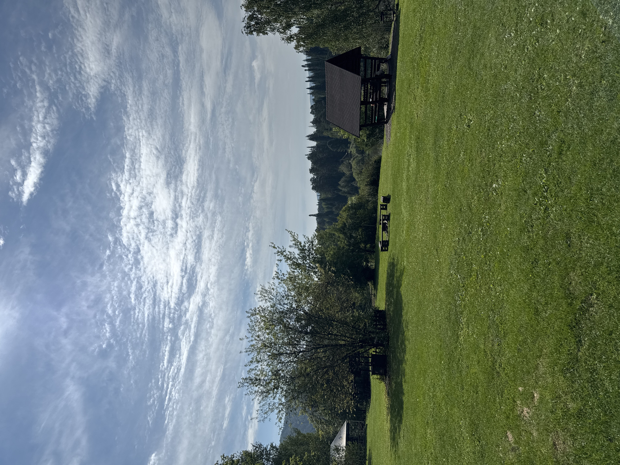 The image depicts a clearing with a well-maintained lawn, bordered by trees and a forested hillside in the background. The sky is partially cloudy with a bright sun peeking through. In the foreground, a vast expanse of green grass dominates the scene. To the left, a small white building with a pitched roof is visible amongst the trees. In the center, a large deciduous tree casts a shadow on the grass, and beneath it, there's a dark, square-shaped object. Further to the right, a dark wooden gazebo with a dark pitched roof stands on a gravel base, with benches and people inside. In the mid-ground, a wooden picnic table with benches is positioned on the grass. To the far right, a single dark wooden bench is placed on the lawn. The overall impression is that of a recreational area or park. The location is identified as Ust-Mana, Russia.