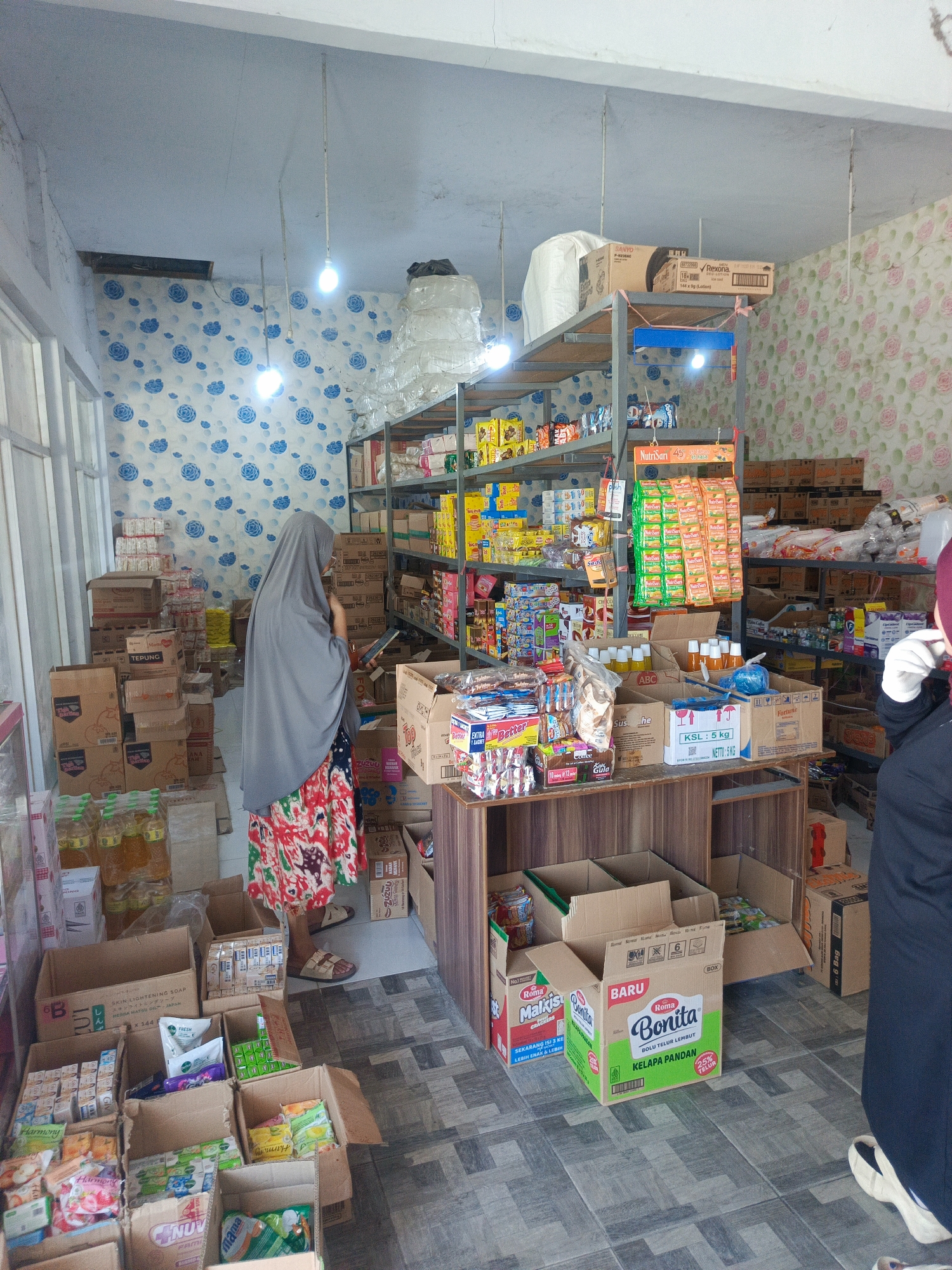 The image depicts the interior of a small retail store, likely a minimarket or warung, located in Tenggela, Indonesia. The store is filled with shelves and stacks of various consumer goods, including packaged food items, drinks, toiletries, and cleaning supplies.
In the center of the frame, a person wearing a grey hijab and a colorful patterned skirt is standing and looking at a mobile phone. They are facing towards the right side of the image. To the right of this person, another individual, partially visible from the side and wearing black clothing and a maroon head covering, is also looking towards the right. This person appears to be wearing white gloves.
The store's interior is characterized by its modest setup. Shelving units, made of metal, are stocked with products arranged in boxes and on display. The walls are decorated with patterned wallpaper, featuring blue floral motifs on one side and pink floral motifs on the other. The floor is tiled in a grey and white checkered pattern. Lighting in the store appears to be provided by overhead fluorescent bulbs.
Various products are identifiable, including bottles of cooking oil, boxes of biscuits or cakes (such as "Bonita" and "Roma Malkist"), soap packages, and other assorted grocery items. The arrangement of goods suggests a typical retail environment where products are organized for sale. Cardboard boxes are also visible, some stacked and some open, indicating stock or recent deliveries. The overall impression is of a well-stocked, albeit small, neighborhood store.