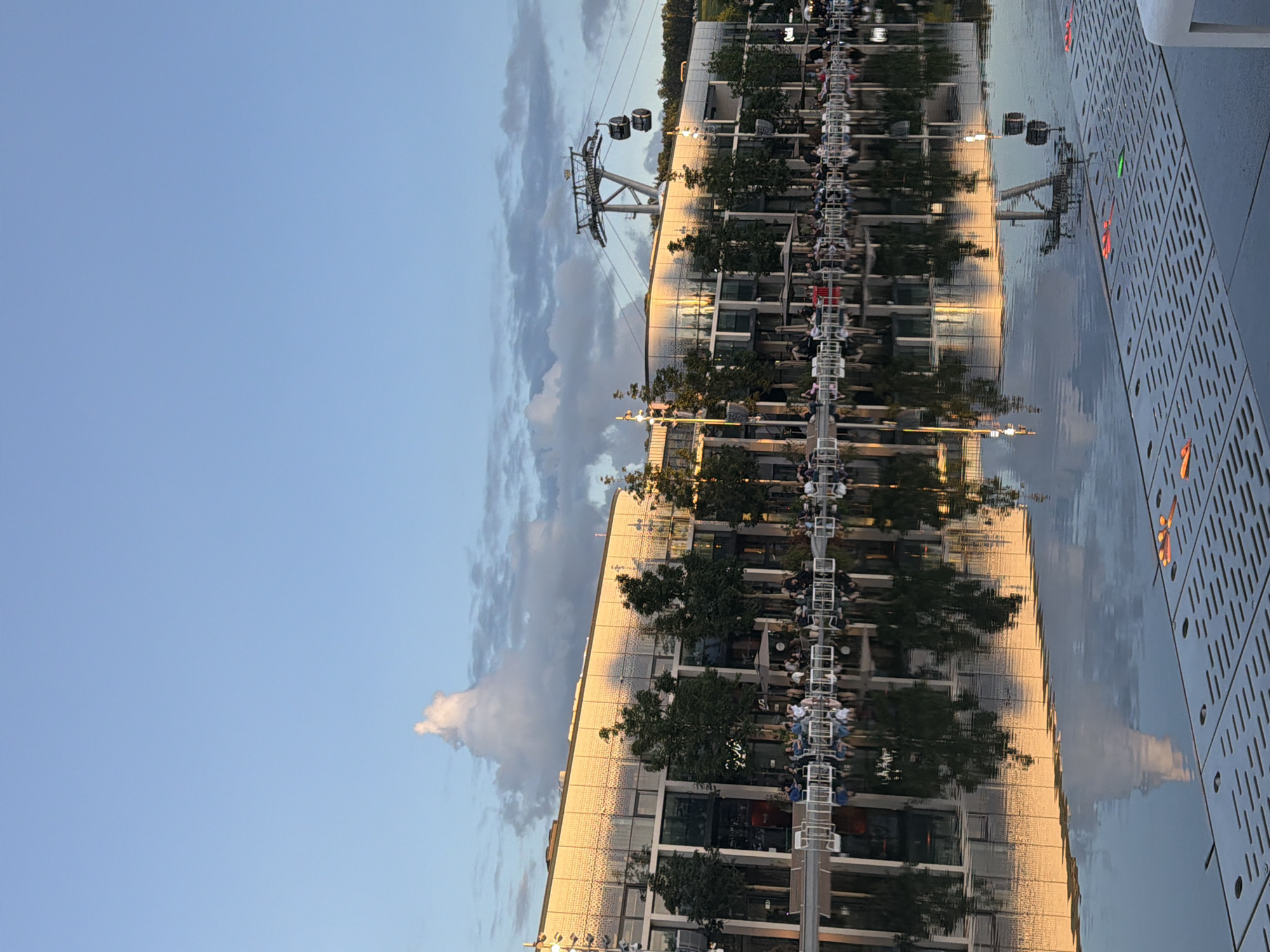 This image depicts an outdoor scene at dusk in an urban area with a reflection pool in the foreground. The setting sun casts a warm golden light on a modern, multi-story building with a patterned facade and large glass windows. Trees are planted along the front of the building, and outdoor seating areas with tables and chairs are occupied by numerous people who appear to be dining or socializing. A cable car system with two cabins is visible against the sky in the upper right portion of the image. The water in the reflection pool mirrors the building, trees, sky, and the cable car structure, creating a symmetrical effect. The sky is a soft blue with scattered clouds, one of which is particularly distinctive, appearing somewhat animal-shaped. The foreground of the reflection pool features a textured grate with some orange and green illuminated elements.