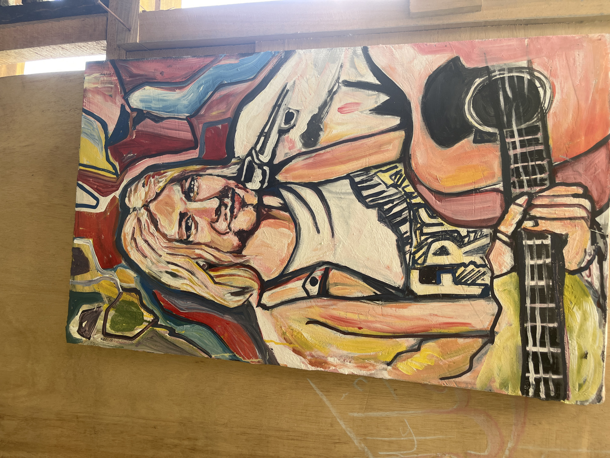 The media file displays a painting, rendered in a vibrant, expressive style with thick black outlines, resting on a light brown wooden surface. The painting depicts a blonde-haired person, likely female, lying horizontally and looking towards the viewer with a slight smile. They are holding or resting with an acoustic guitar.
The person in the painting has light skin, large eyes, and is wearing a white top. Abstract patterns, possibly tattoos or designs on the clothing, are visible on their left arm and shoulder area. The acoustic guitar, colored predominantly in shades of reddish-pink for its body, features a black soundhole and fretboard with visible strings. The person's right hand appears to be positioned on the neck of the guitar, while the left hand is near the body.
The background of the painting is composed of abstract, angular shapes in a palette of red, blue, yellow, green, and brown, creating a dynamic and colorful backdrop for the central figure. The brushstrokes are visible, giving the painting a textured appearance.
The photograph itself is taken in bright conditions, with a strong light source entering from the top, causing some glare. The wooden surface on which the painting is displayed shows a natural grain, and behind the painting, wooden beams or planks are visible, suggesting an indoor or covered outdoor setting like a studio or workshop. There is no visible text within the painting or the surrounding environment in the image.