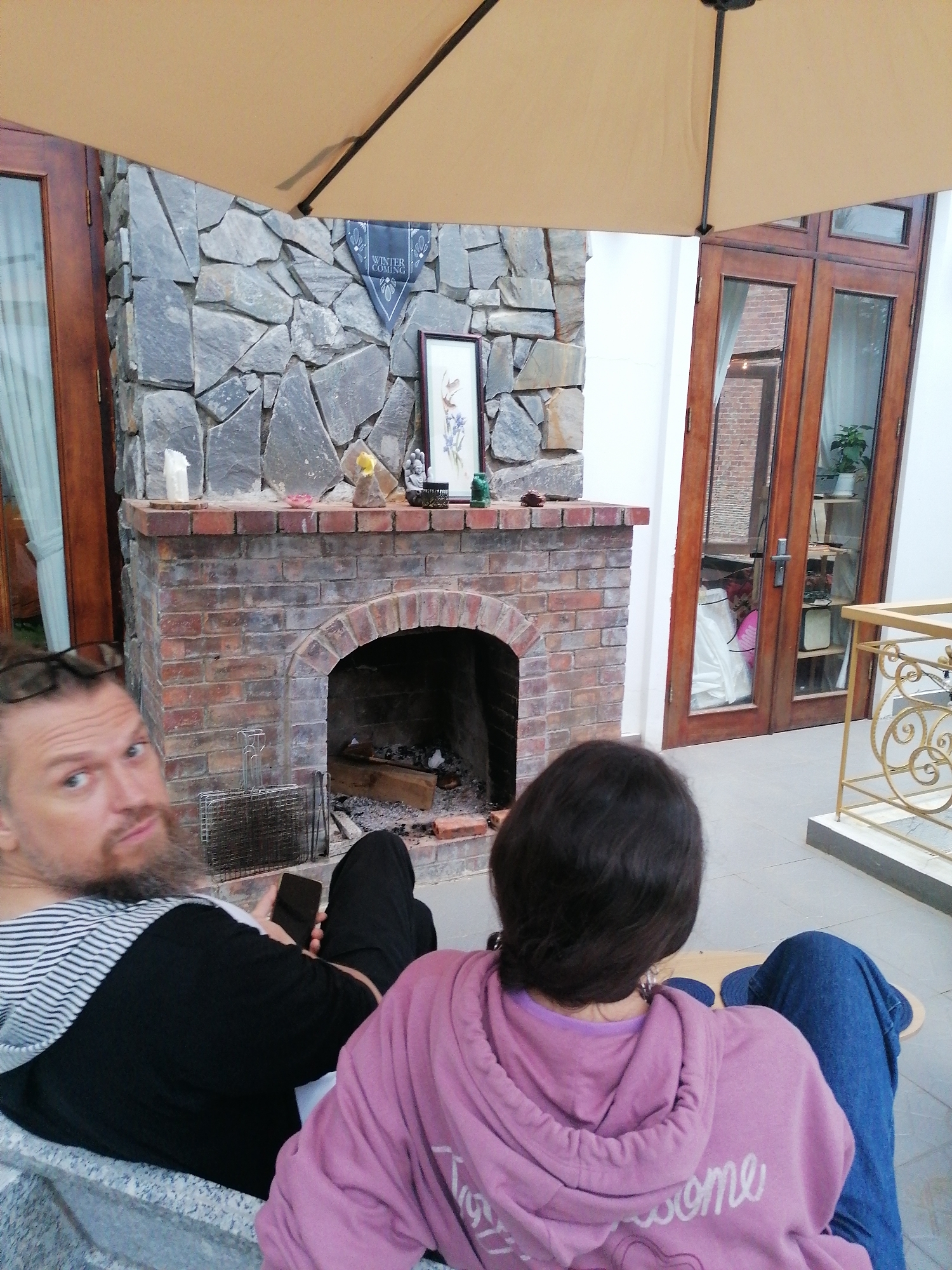 The image captures an outdoor or semi-outdoor patio scene in Lạc Dương, Vietnam, featuring two individuals relaxing by a rustic fireplace. In the foreground, a man with a beard and eyeglasses resting on his head looks directly at the camera with a neutral expression. He is wearing a black long-sleeved shirt with a striped hood and holds a smartphone. Next to him, a woman is seen from behind, wearing a purple hoodie with the word "awesome" partially visible in white text on the back, and blue jeans.
A prominent fireplace forms the central background, constructed with a base of red bricks and an upper section of irregularly shaped grey stones. Its arched firebox contains burnt wood and a metal grill grate. The mantelpiece is adorned with decorative items including a white candle, small figurines, and a framed picture. Above, a dark banner displays the text "WINTER IS COMING".
Flanking the fireplace are wooden doors with glass panels, suggesting entry to an interior space. The doors on the right reflect a brick-walled room with a potted plant. A large, beige patio umbrella partially shades the scene from above. To the far right, a golden, ornate metal railing defines the edge of the patio. The scene conveys a relaxed daytime atmosphere in a cozy, outdoor setting.