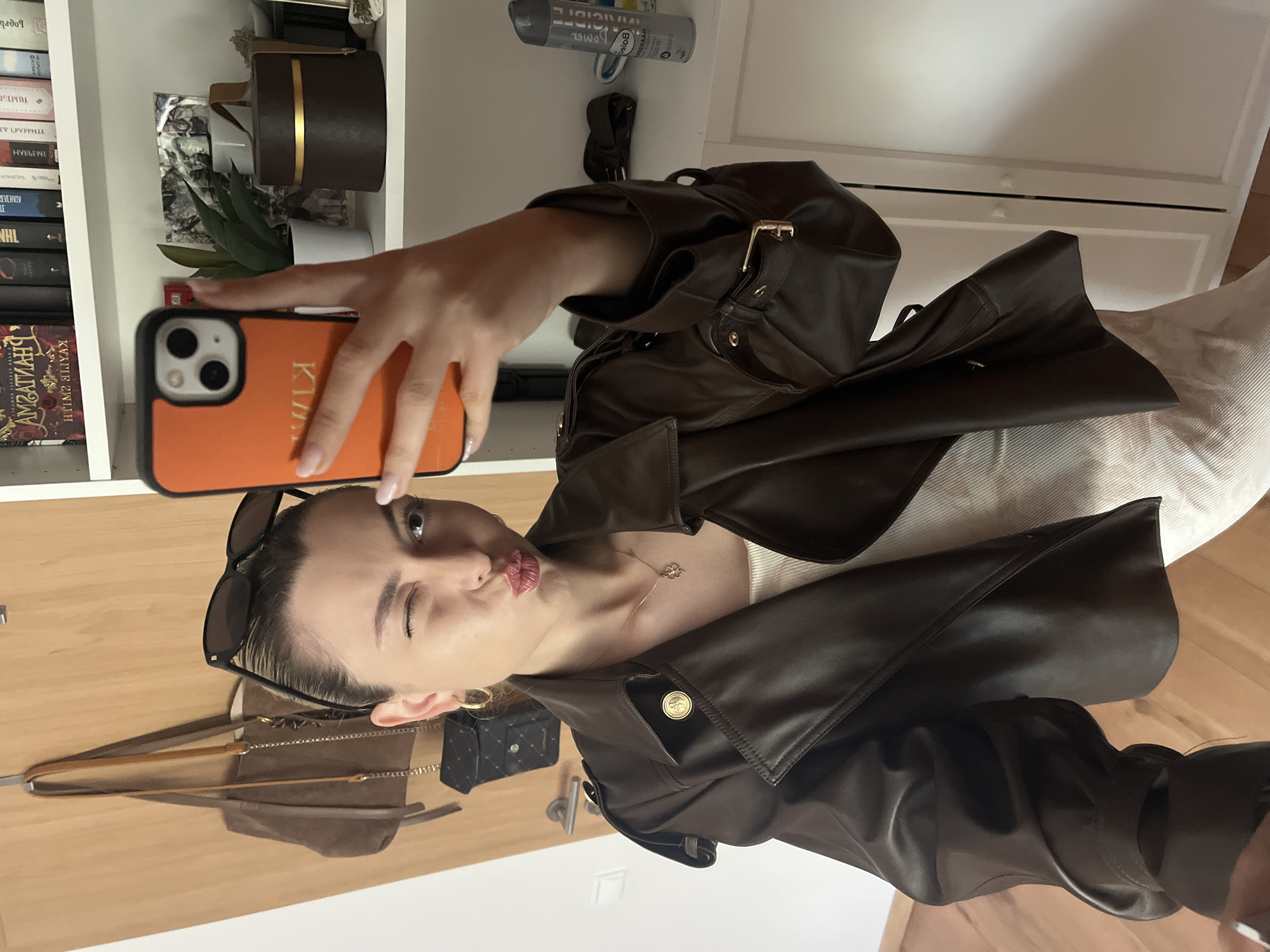 A young woman is taking a selfie in what appears to be a bedroom. She is wearing a dark brown leather jacket and has sunglasses perched on her head. She is holding an orange phone with a clear case. Her mouth is puckered into a kiss and she is winking one eye. In the background is a shelf with books and decorative items. A handbag hangs on the door behind her. The location appears to be Unknown, Unknown.
