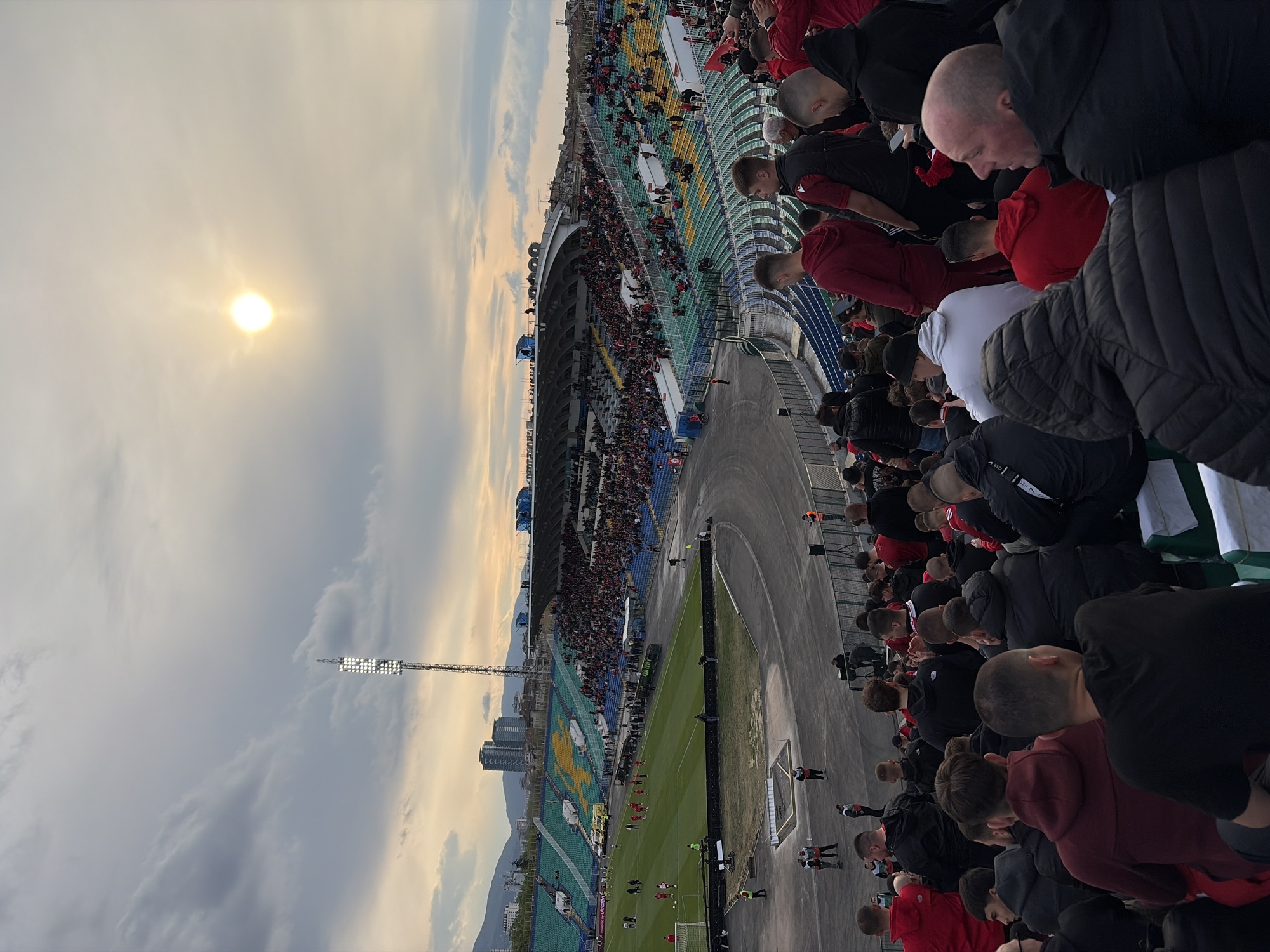 This panoramic image captures a vibrant football (soccer) stadium scene from an elevated spectator's perspective. The sky, bathed in the soft glow of a late afternoon or early evening sun, dominates the upper-left, casting a warm light over scattered clouds.
The stadium features multi-tiered stands curving around the pitch, filled with numerous spectators. The seats are a mix of green, yellow, and blue, with a large yellow symbol, possibly a letter or club emblem, formed by yellow seats on one side. In the foreground and midground, many spectators are visible, predominantly wearing dark clothing like black jackets and hoodies, interspersed with some red shirts. Their attention is directed towards the field below.
The green football pitch occupies the center-lower portion of the frame, encircled by a grey track. A few figures, likely players or staff, are visible on the field, suggesting a match or event is underway. Part of a goalpost can be seen at the edge of the pitch.
Beyond the stadium, a tall floodlight tower stands against the cloudy sky. In the far distance, city buildings rise above the horizon, with mountains partially visible behind them, consistent with a location like Sofia, Bulgaria. The weather is partly cloudy, and the sun appears somewhat hazy. No significant text is clearly discernible, apart from the large yellow symbol on the seats.