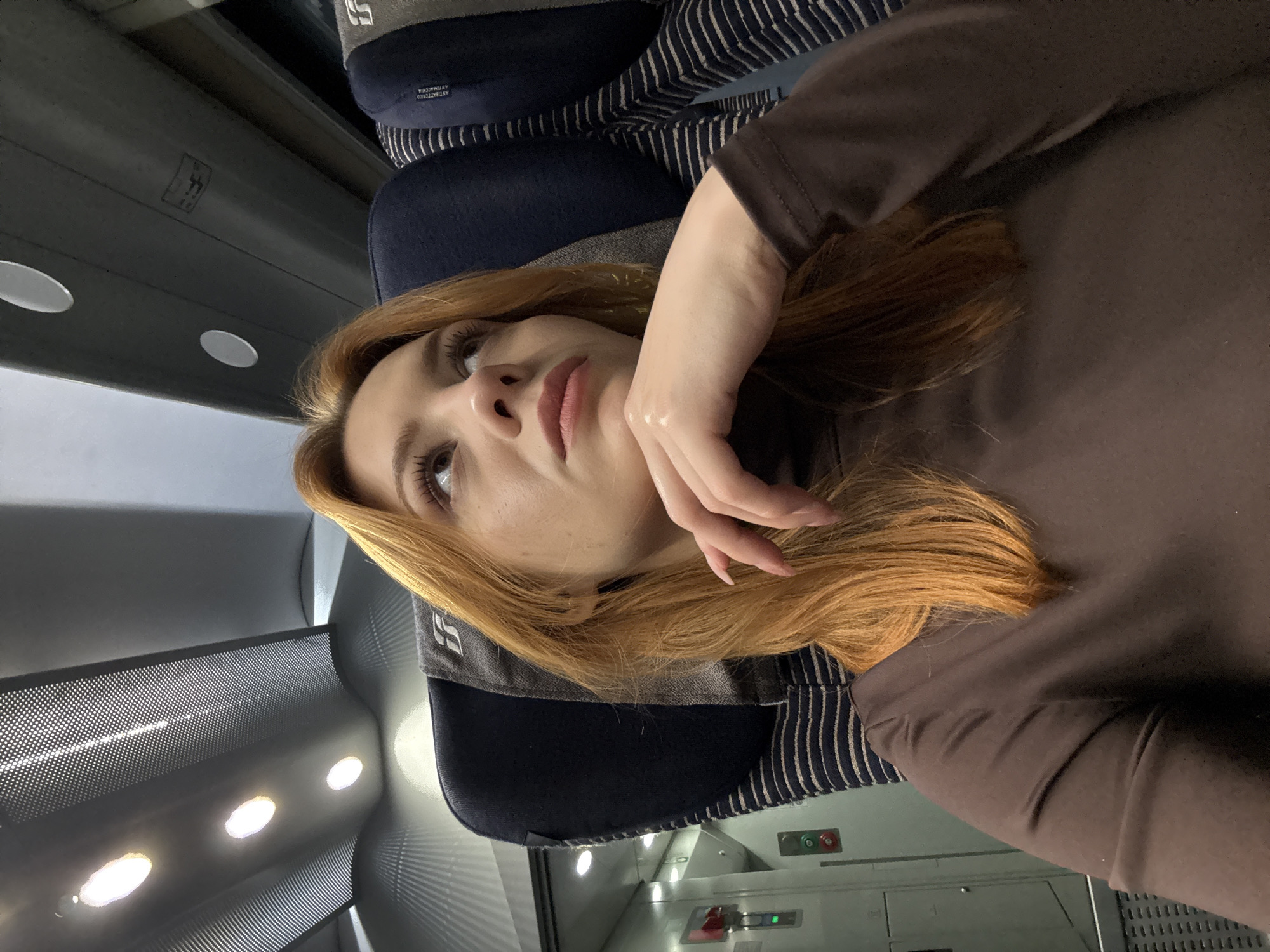 A woman is lying down in a train car. She has light skin, brown eyes, and long, reddish-blonde hair. She is wearing a brown long-sleeved shirt. Her left hand is raised near her face. She appears to be on a train with blue and grey striped seats. The train car interior is visible with overhead lighting and air conditioning vents. The specific location in Borgio Verezzi, Italy cannot be determined from the image.