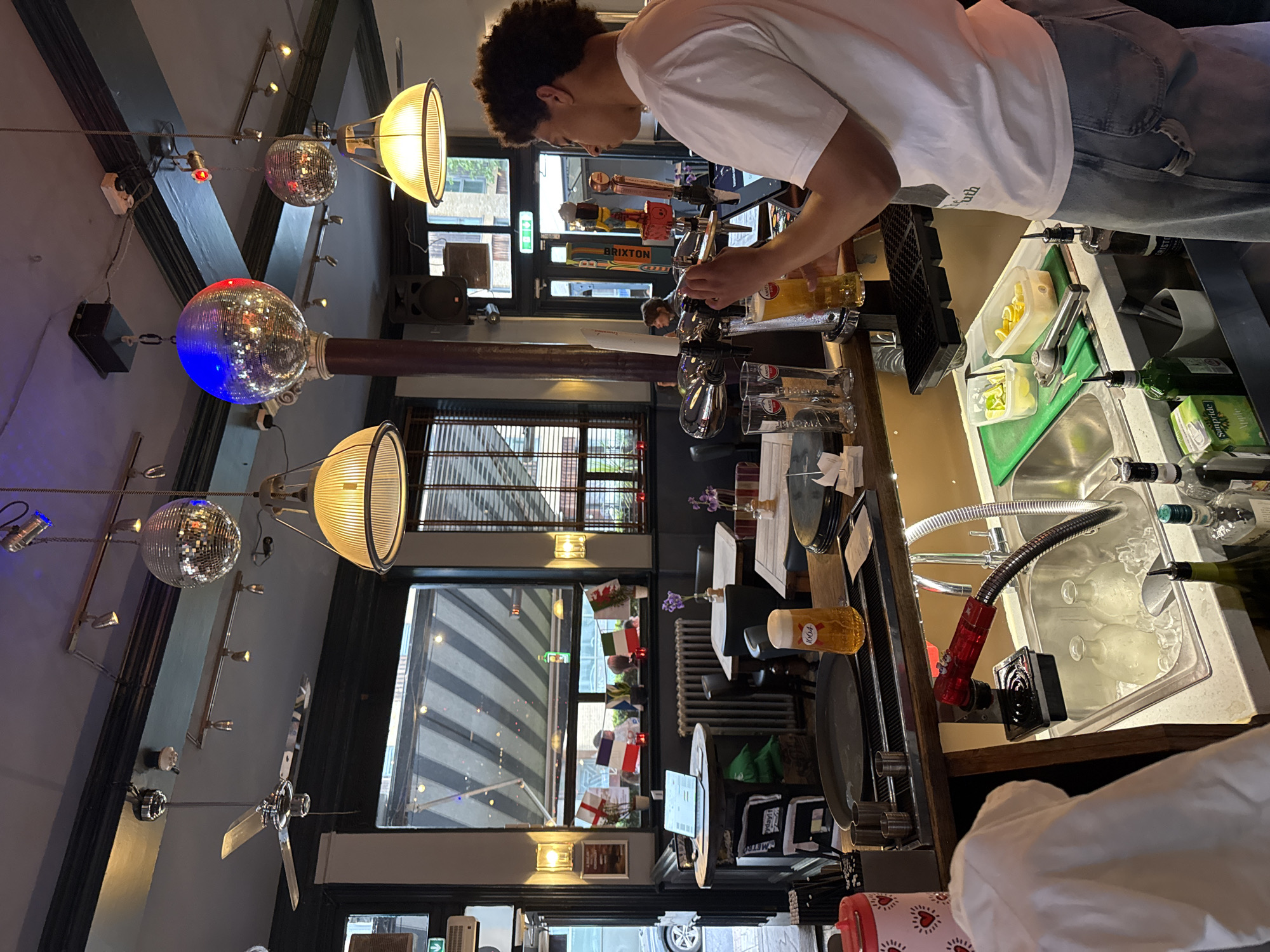An indoor shot of a pub or bar, possibly in London, United Kingdom. A barman is seen filling a glass with beer from a tap behind the bar. Decorations include disco balls and lights hanging from the ceiling. Various glasses, bottles, and bar equipment are present on and behind the counter. Through the window, a view of the street is visible, including various flags displayed.