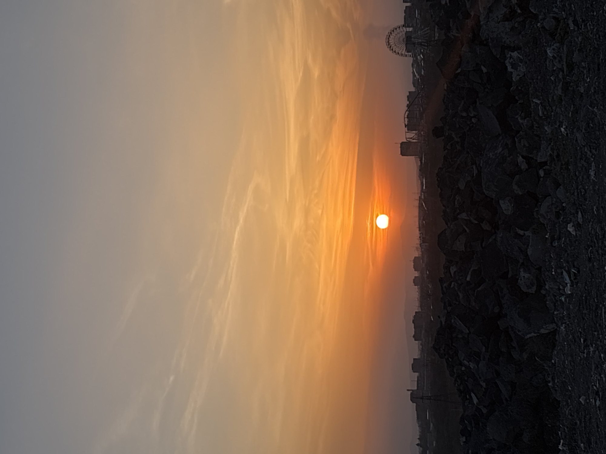 The image depicts a sunset in Yerevan, Armenia. The sun is setting on the right side of the frame, casting an orange glow across the sky. The sky features wispy clouds. In the distance, a Ferris wheel and other structures are silhouetted against the setting sun. The foreground shows a dark, rocky terrain.