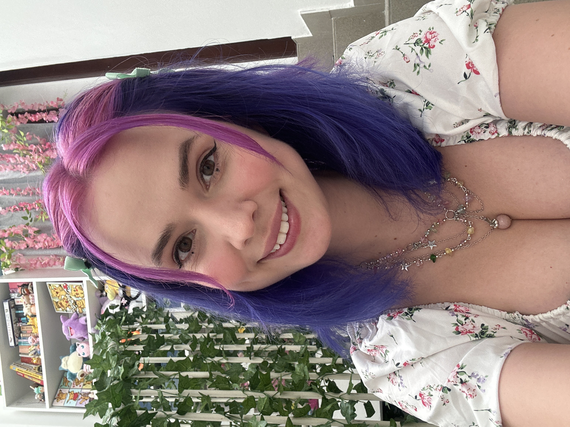A young woman with dyed pink and blue hair smiles towards the viewer. She wears a floral-print dress and a layered necklace. Behind her is a white bookshelf filled with various items, including what appears to be plush toys and books. A trellis covered in artificial ivy stands in front of the bookshelf. A string of artificial pink flowers is also visible behind her. The scene takes place indoors with natural light. There is no indication as to whether or not the image was taken in City of Zagreb, Croatia.