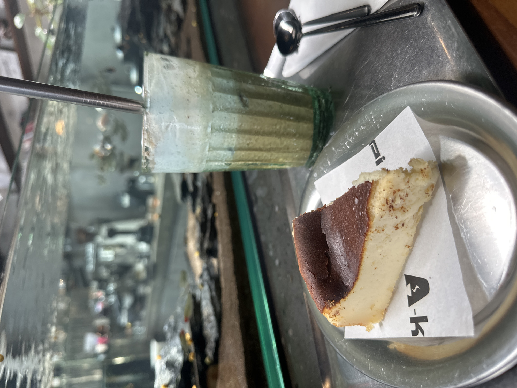 A slice of Basque cheesecake rests on a silver plate lined with a white paper napkin featuring a black logo. Next to it, a tall glass contains a layered drink, possibly a matcha latte or a blended beverage, with a metal straw inserted. The background is out of focus, showing what appears to be a busy cafe or restaurant environment with glass partitions, possibly a display of food or drinks, and patrons in the distance. The overall scene suggests a pleasant cafe experience. While the image is visually rich, it does not contain any information that would place it in Da Nang, Vietnam, or any specific city.