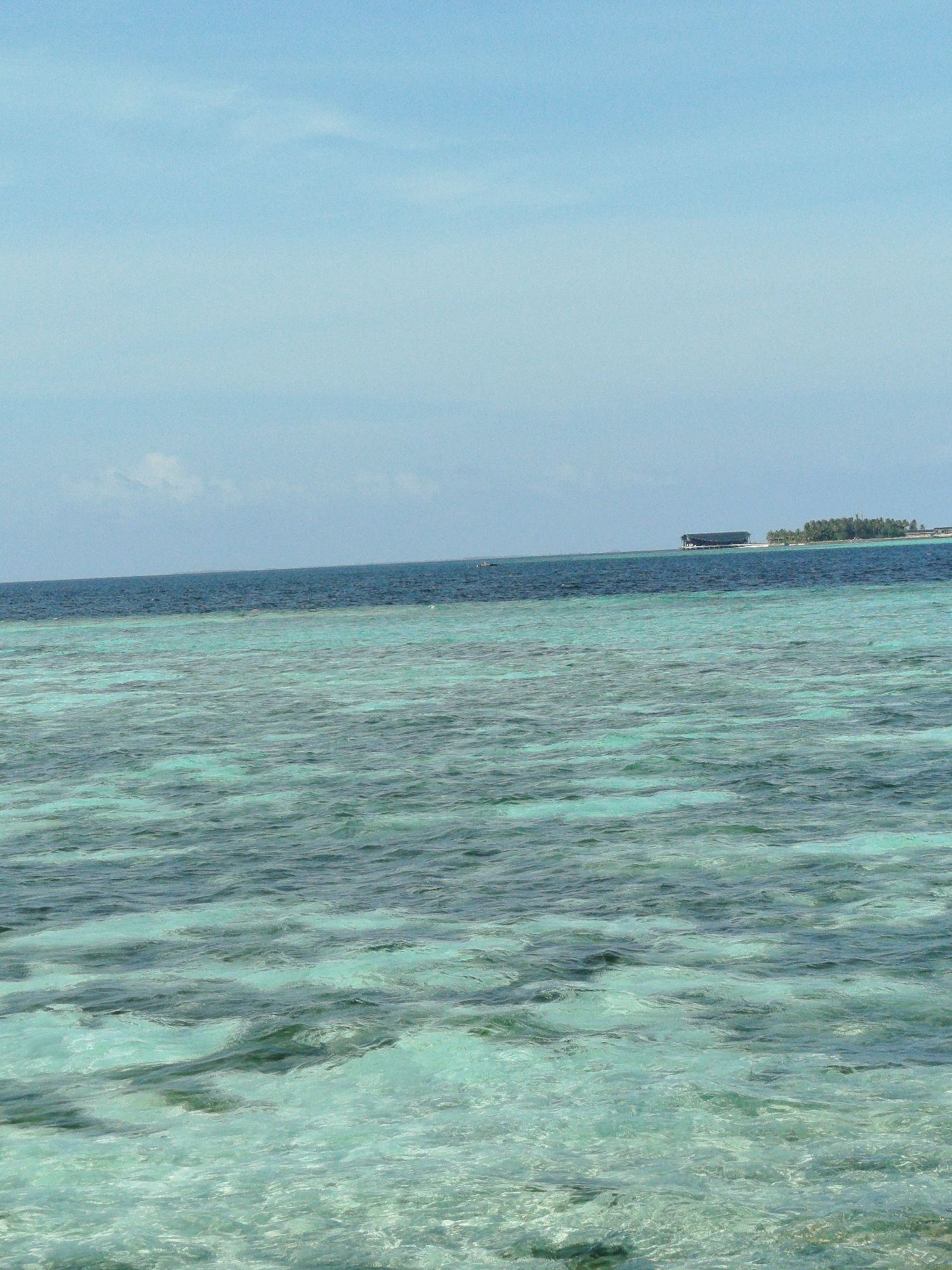 A wide shot captures a section of the ocean near Hinnavaru, Maldives, under a pale blue sky. The water is divided into two distinct layers: the foreground is a shallow, turquoise expanse with visible sandbanks beneath the surface, while the background displays a deeper, darker blue ocean. In the distance, a narrow strip of land with lush green vegetation is visible, with a long, dark structure, possibly a resort or a floating platform, situated on the water near the island. The sea surface is rippled, indicating gentle waves. There are no people or animals explicitly visible in the frame. The overall impression is one of a serene tropical seascape.