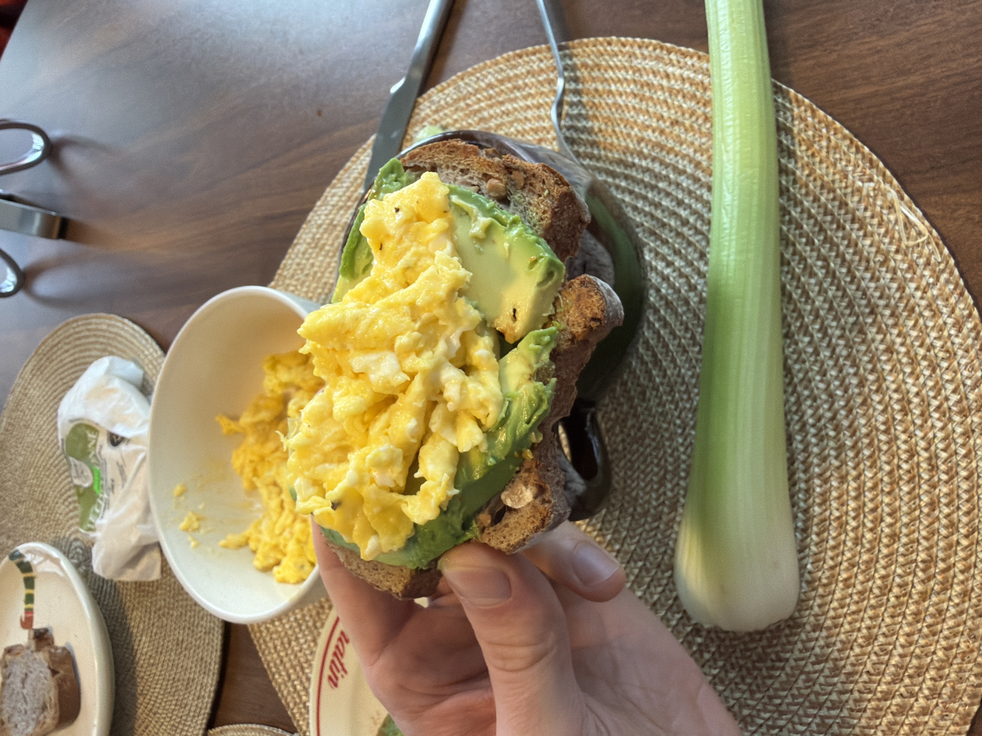 The image depicts a hand holding a sandwich made of whole wheat bread, avocado slices, and scrambled eggs. To the left of the sandwich is a small bowl containing more scrambled eggs, and a small plate with a piece of bread. To the right of the sandwich is a whole celery stalk. A knife and fork are on top of the tan braided placemat. The location is likely indoors on a wooden table. There is no discernible information in the image to suggest it is in Montreal, Canada.