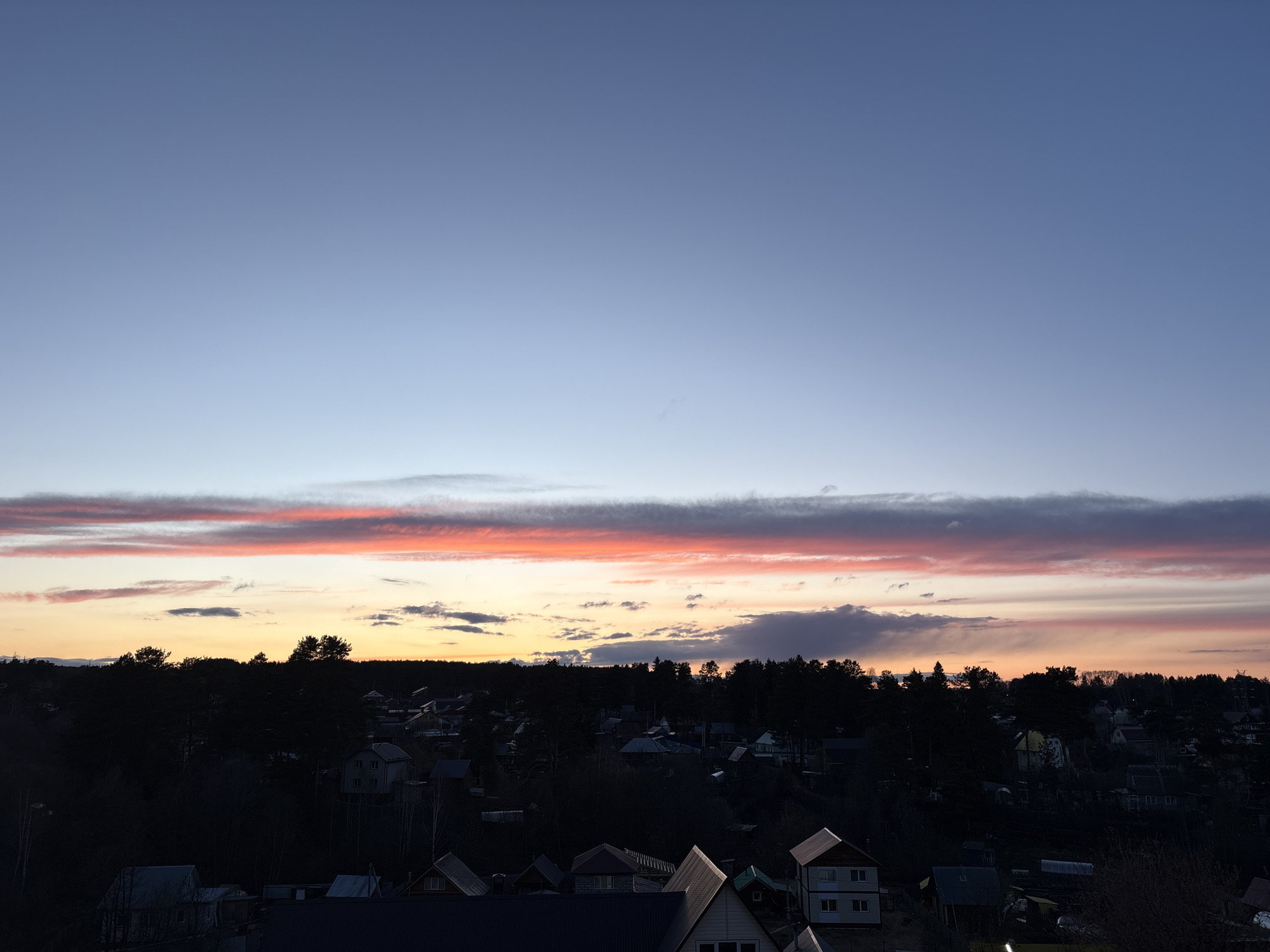 This media file captures a serene evening landscape in Chaikovsky, Russia, characterized by a vibrant sunset sky over a dark forest and a residential area. The upper two-thirds of the image display a gradient sky, transitioning from a deep twilight blue at the top to lighter blues near the horizon. A prominent horizontal band of clouds, illuminated by the setting sun, spans across the middle of the sky with striking hues of orange, pink, and red, intermingled with darker grey cloud formations.
Below the colorful sky, a dense line of trees, primarily coniferous, forms a dark silhouette along the horizon, suggesting a substantial forest. Nestled beneath this forest and extending into the foreground are numerous houses, forming a village or suburban settlement. These structures appear largely in shadow due to the low light, with only the shapes of their roofs and some outlines of walls discernible. The architecture of the visible houses, with their sloped roofs and varying sizes, is typical of rural or dacha-style dwellings.
The scene depicts a peaceful moment at sunset or just after, with the ground details obscured by darkness, highlighting the dramatic sky. The weather appears to be partly cloudy, allowing both clear sky and colorful cloud formations to be visible. There is no discernible activity taking place, nor is any visible text present in the image. The overall impression is one of quiet natural beauty at dusk.