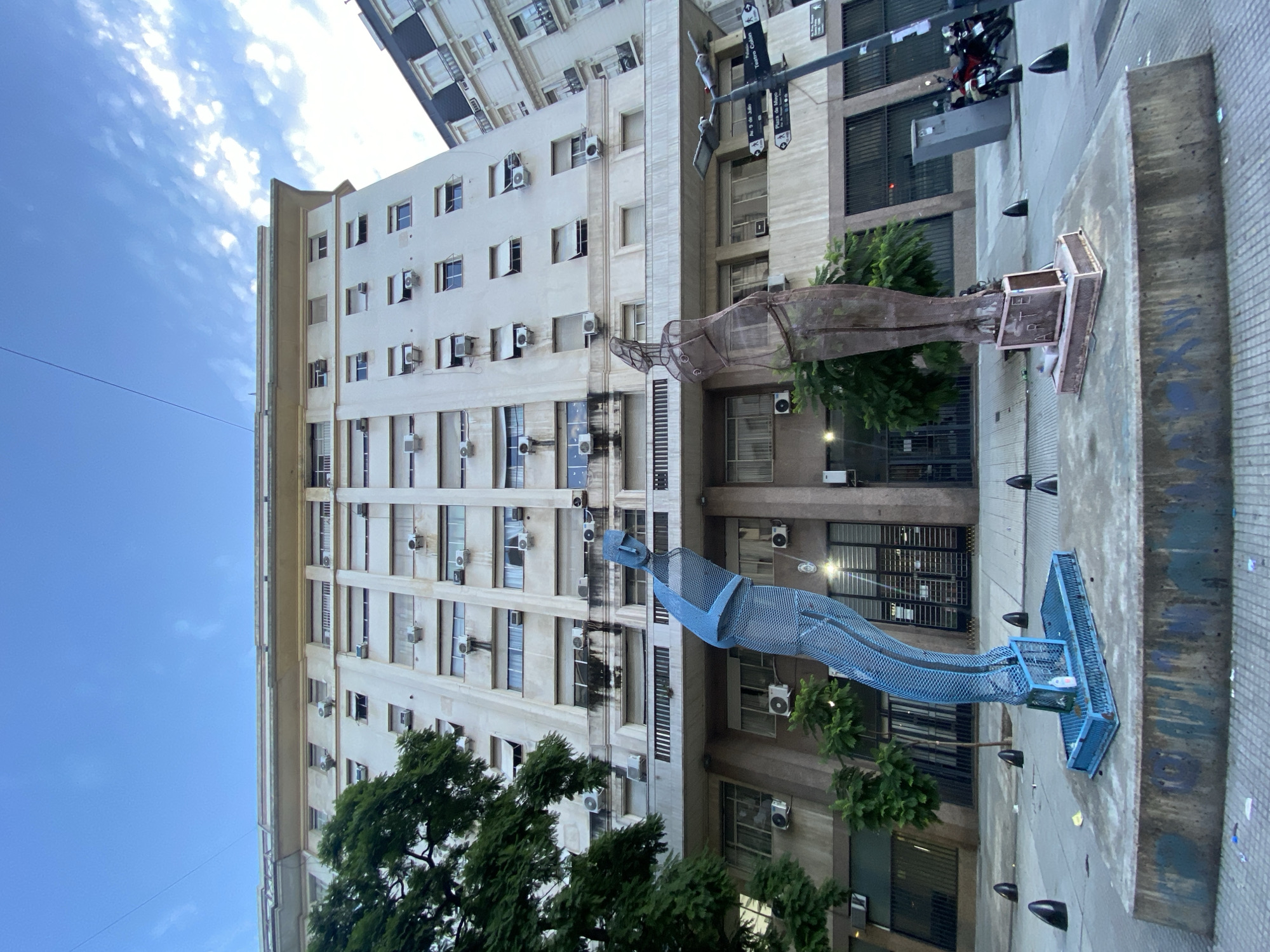 The image shows an urban scene in Buenos Aires, Argentina. Two large statues, one blue and one brown, stand in front of a tall building. The blue statue appears to be a person lying down, constructed of mesh, while the brown statue is an abstract human form. The building has multiple stories with rows of windows and numerous air conditioning units. Streetlights and trees are present, and the sky is partially visible. The scene likely depicts an outdoor public art installation in a city setting.