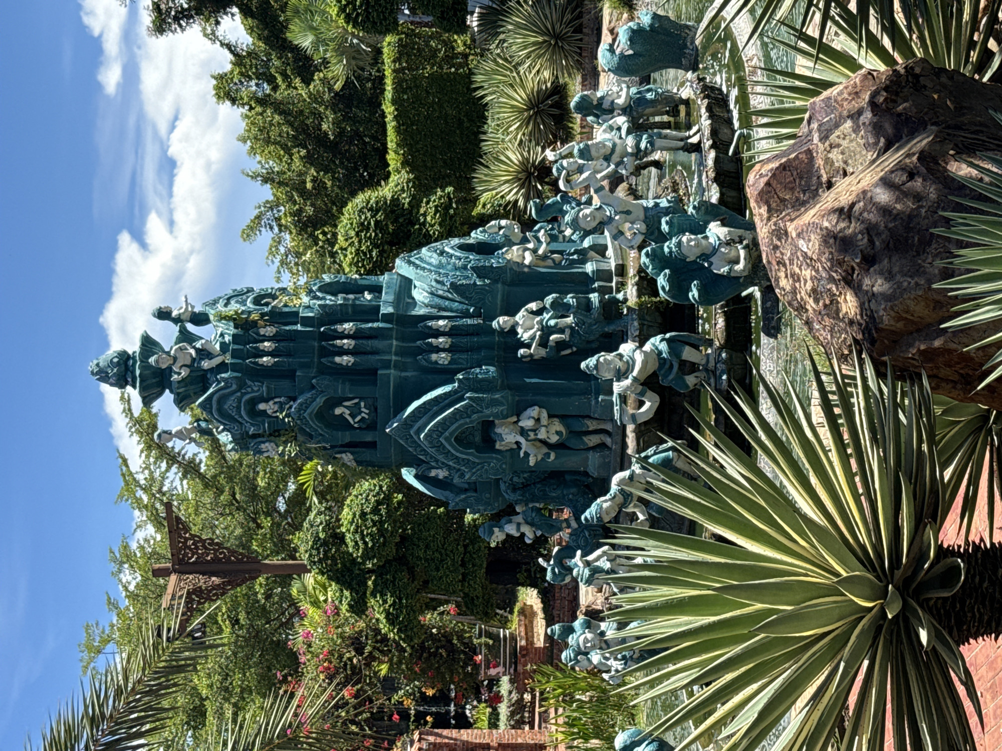 The image shows a multi-tiered turquoise-colored monument adorned with figures, possibly depicting a scene from mythology or local folklore. This structure is situated within a landscaped garden, identifiable by various plant species, including palm trees, agave plants, and other greenery. A large rock sits adjacent to the monument, adding to the natural elements of the setting. The sky is visible in the background, indicating it is daytime. The location is Bang Pu Subdistrict Municipality, Thailand.