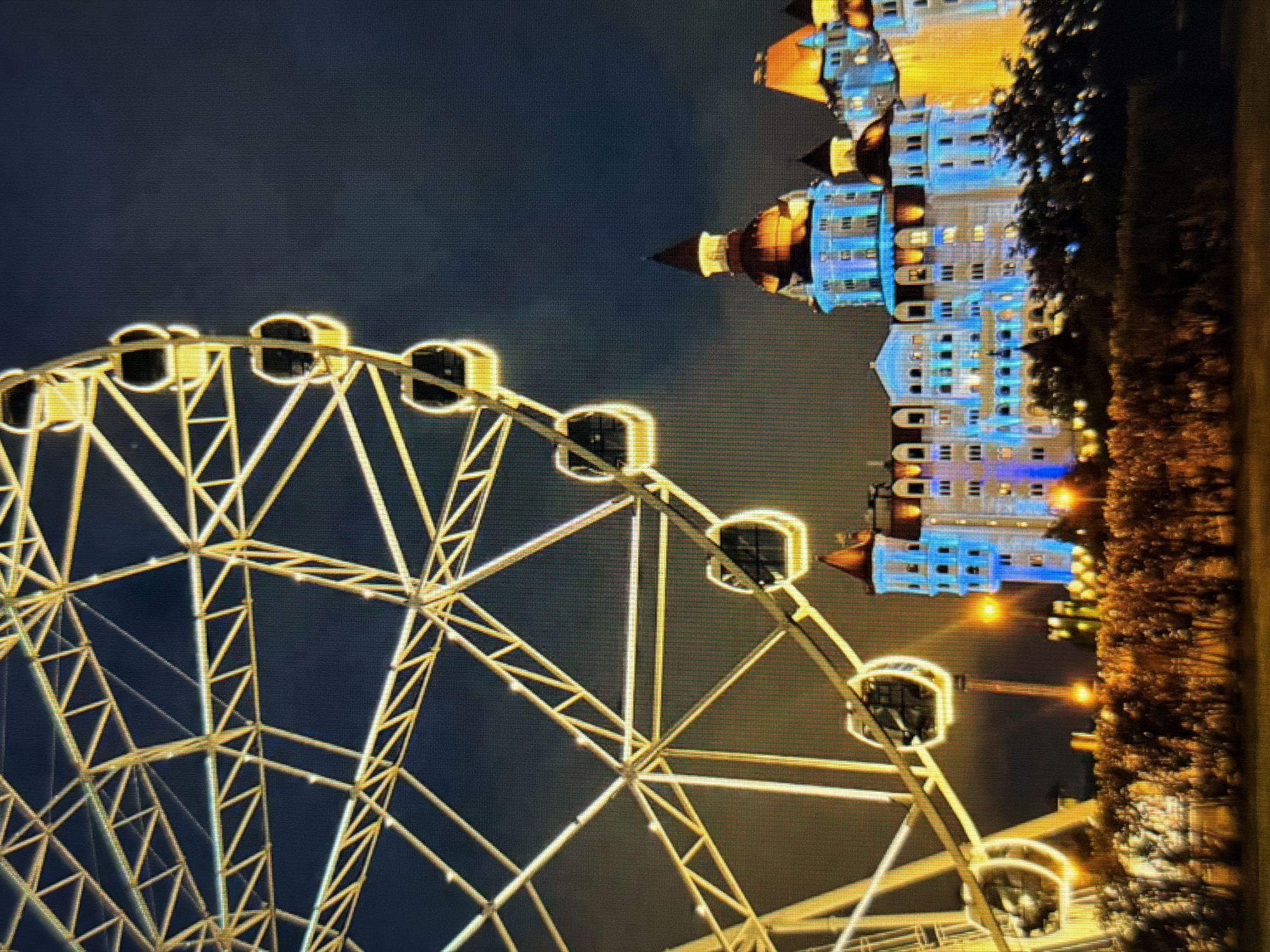 This image depicts a nighttime scene in Sirius, Russia, featuring a large, illuminated Ferris wheel in the foreground. The Ferris wheel, constructed with a white metal frame, is adorned with yellow lights that highlight its structure and circular gondolas. Behind the Ferris wheel, a brightly lit building complex with a fairytale-like architectural style is visible. The buildings are illuminated with blue and yellow lights, accentuating their numerous windows, turrets, and domed roofs. A line of illuminated trees and bushes forms a border between the foreground and the building complex, suggesting a park or landscaped area. The sky is dark, indicating nighttime, with some hints of clouds. The overall impression is one of a festive or amusement park setting.